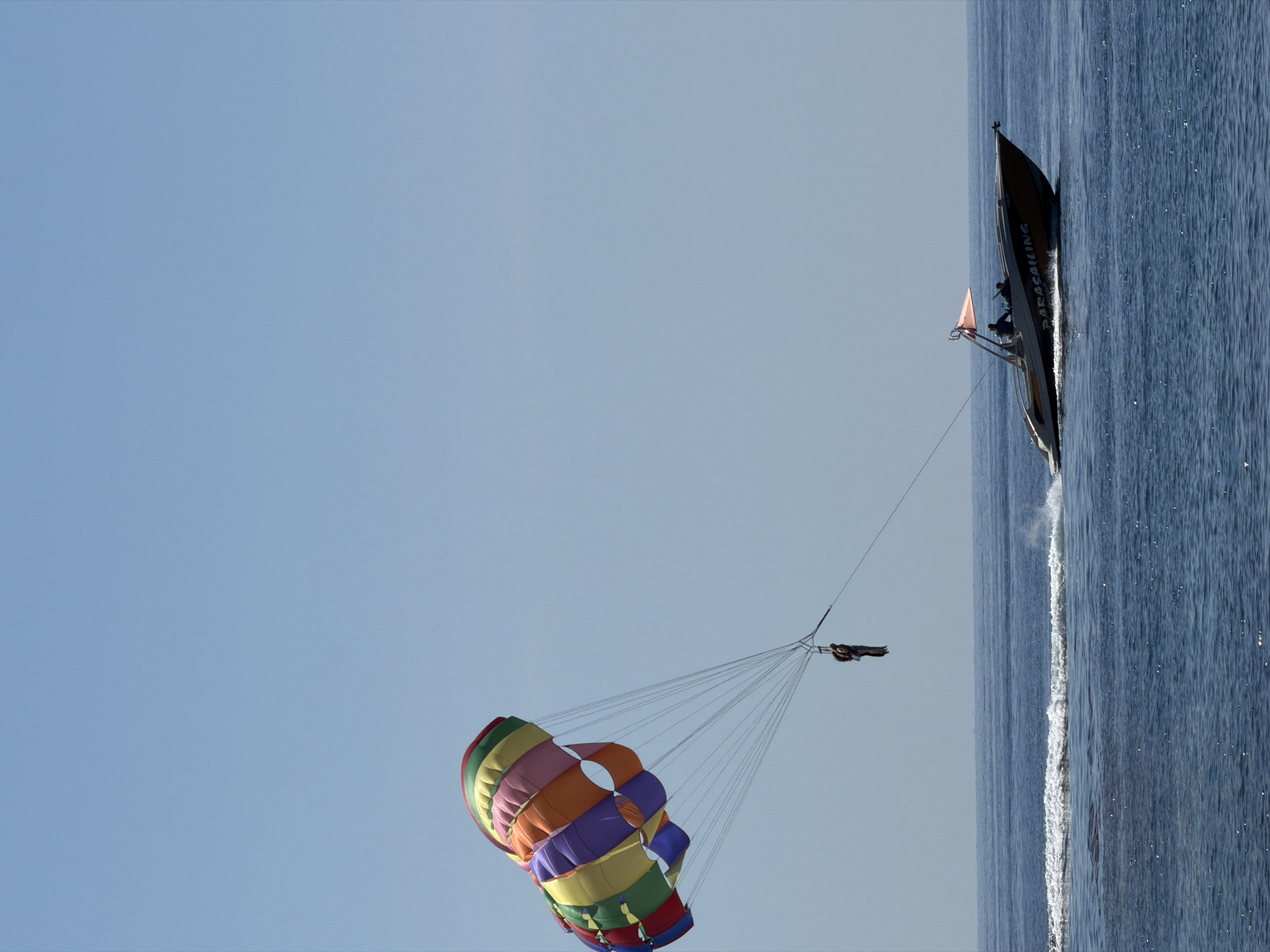 This image captures a parasailing operation in the sea near Budva, Montenegro. A large, multicolored parasail, composed of segments in shades of red, orange, yellow, green, purple, and blue, is airborne against a clear, light blue sky. Suspended beneath the parasail by multiple lines is a harness containing one or two individuals, visible as dark silhouettes. A long tow rope extends from this harness to a motorboat navigating the dark blue water below. The boat, a parasailing vessel, features a dark hull with lighter upper sections and the word "PARASAILING" visible on its side. An orange fabric canopy or flag is affixed to a structure at the boat's stern. At least one person, a dark silhouette, is operating the boat, which is in motion and creating a distinct white wake behind it. The sea surface exhibits minor ripples.