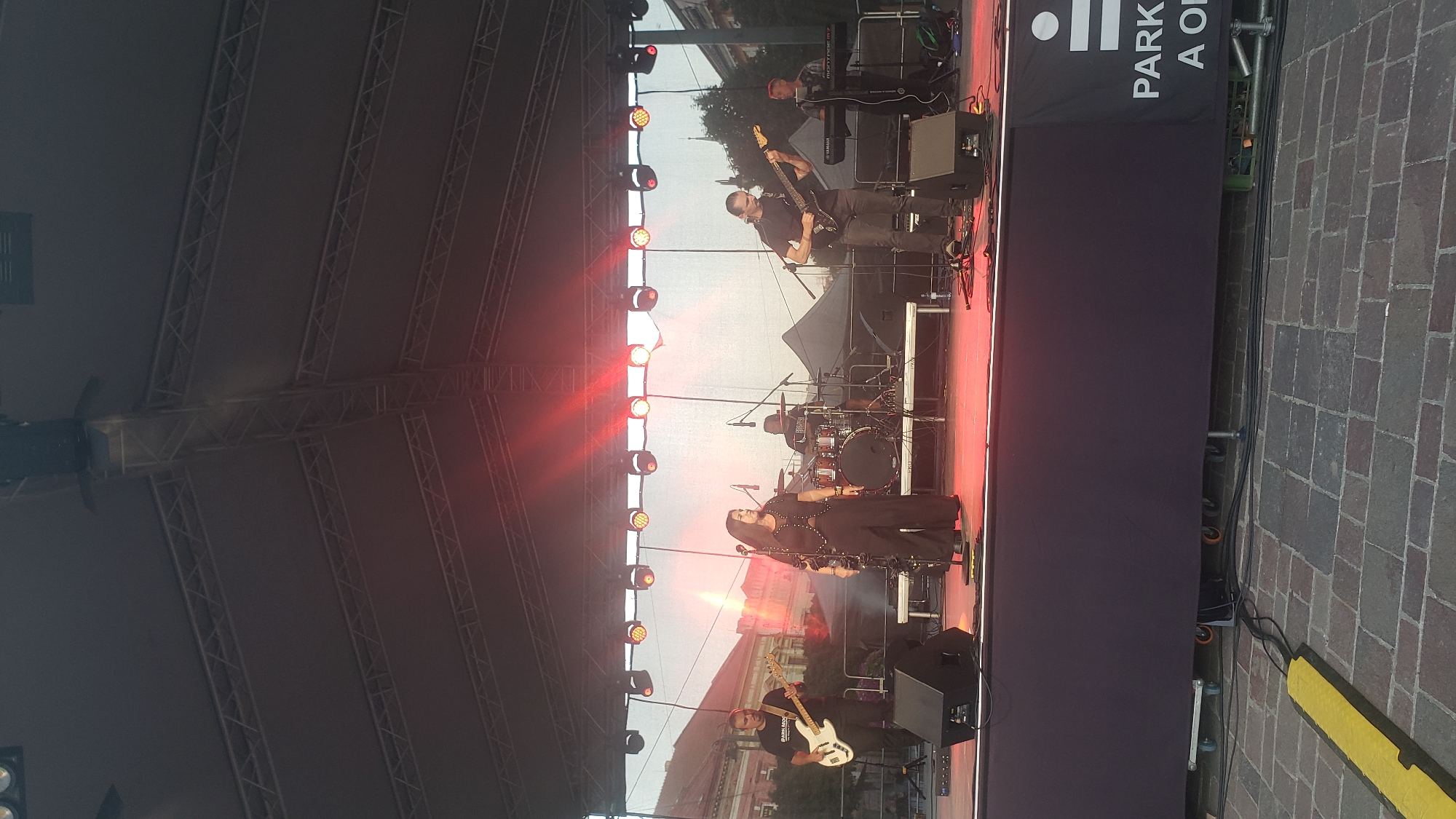 A band performs on an outdoor stage at night, with red stage lights illuminating the performers and the area behind them. The stage is covered by a metal truss roof structure with multiple lights. On the left side of the stage, a man in a black t-shirt plays a white bass guitar. Beside him, a woman in black clothing stands with her hands clasped. Behind her, a drummer is seated at a drum kit. To the right of the drummer, a man with long hair plays a black electric guitar. Further to the right, another man is seated at a keyboard. In front of the stage, a black scrim banner with the text "PARK" and "A O" is visible. The stage is set up on a cobblestone ground. The background shows buildings and trees, suggesting an urban setting. The lighting and the presence of a stage indicate a concert or festival event. This event is taking place at the square of St. Nicholas (Slovenská sporiteľňa) in Prešov, Slovakia, as evidenced by the buildings visible in the background, which are typical of the city's historic center.
