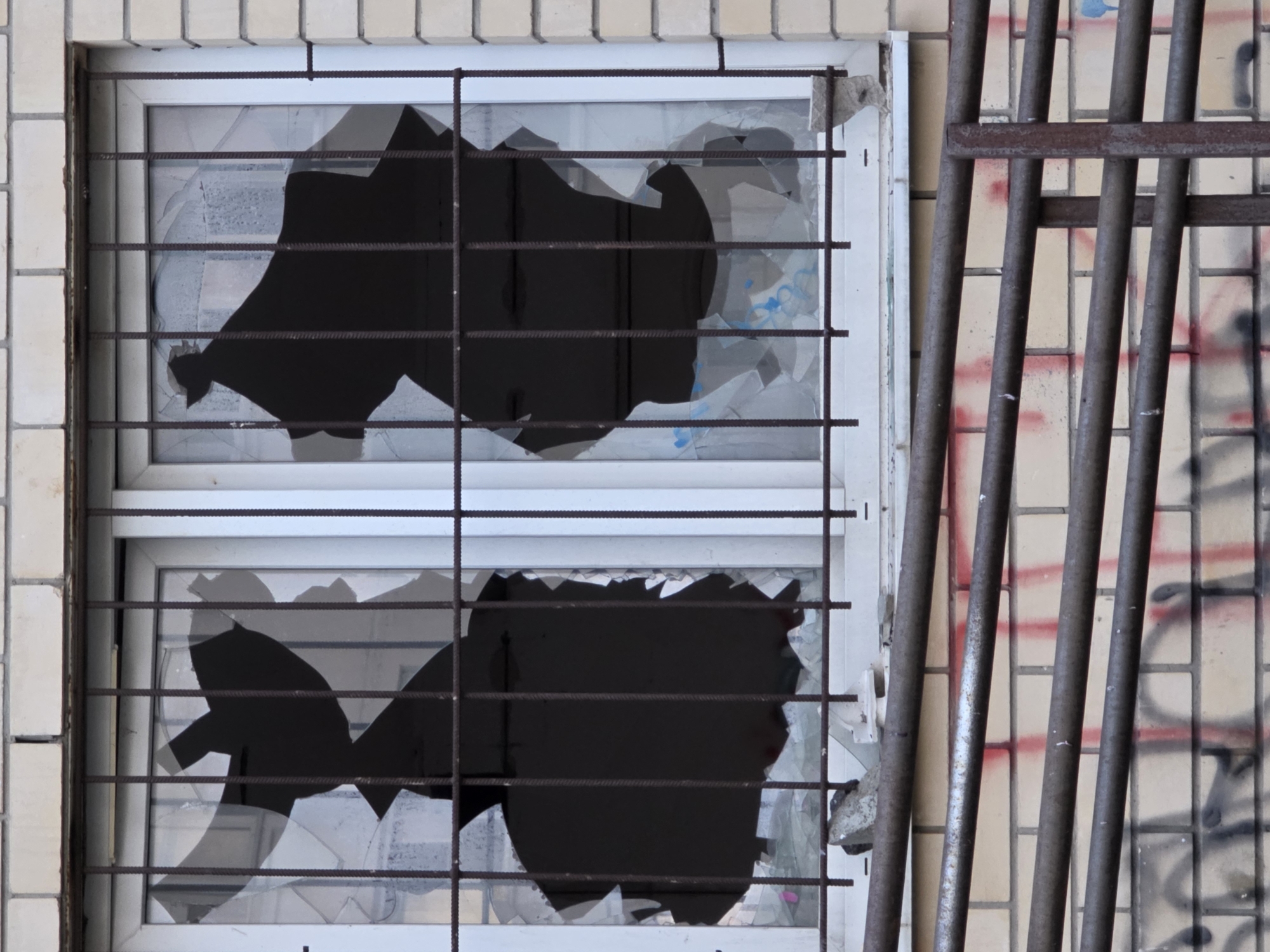 The image shows a close-up of a broken window on a brick building, with metal bars covering it. The glass is shattered into large pieces, creating black voids where the transparency is lost. The window is part of a larger building facade made of light-colored bricks. Below the window, there is a metal railing structure, and the brick wall underneath it is covered in graffiti. The overall scene suggests vandalism or damage to the property. The image does not provide any explicit visual cues to pinpoint its exact location within Rostov-on-Don, Russia.