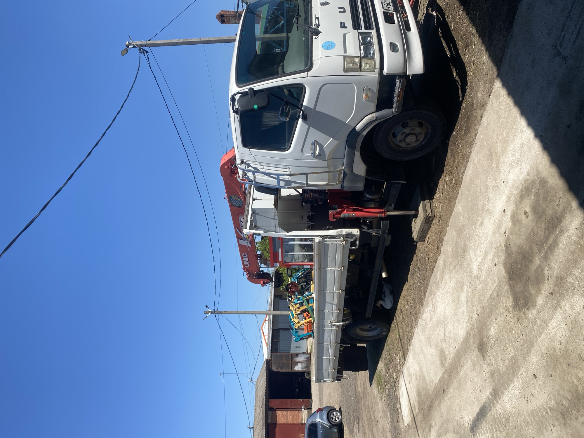 A white, right-hand drive Fuso truck with a red UNIC crane is parked in the city of Neftyanik, Russia. The truck is adjacent to a concrete surface and a weathered building with red and brown walls. Power lines stretch across a clear, blue sky. In the truck's bed, various pieces of construction equipment are visible. A silver passenger car is parked further down the road.