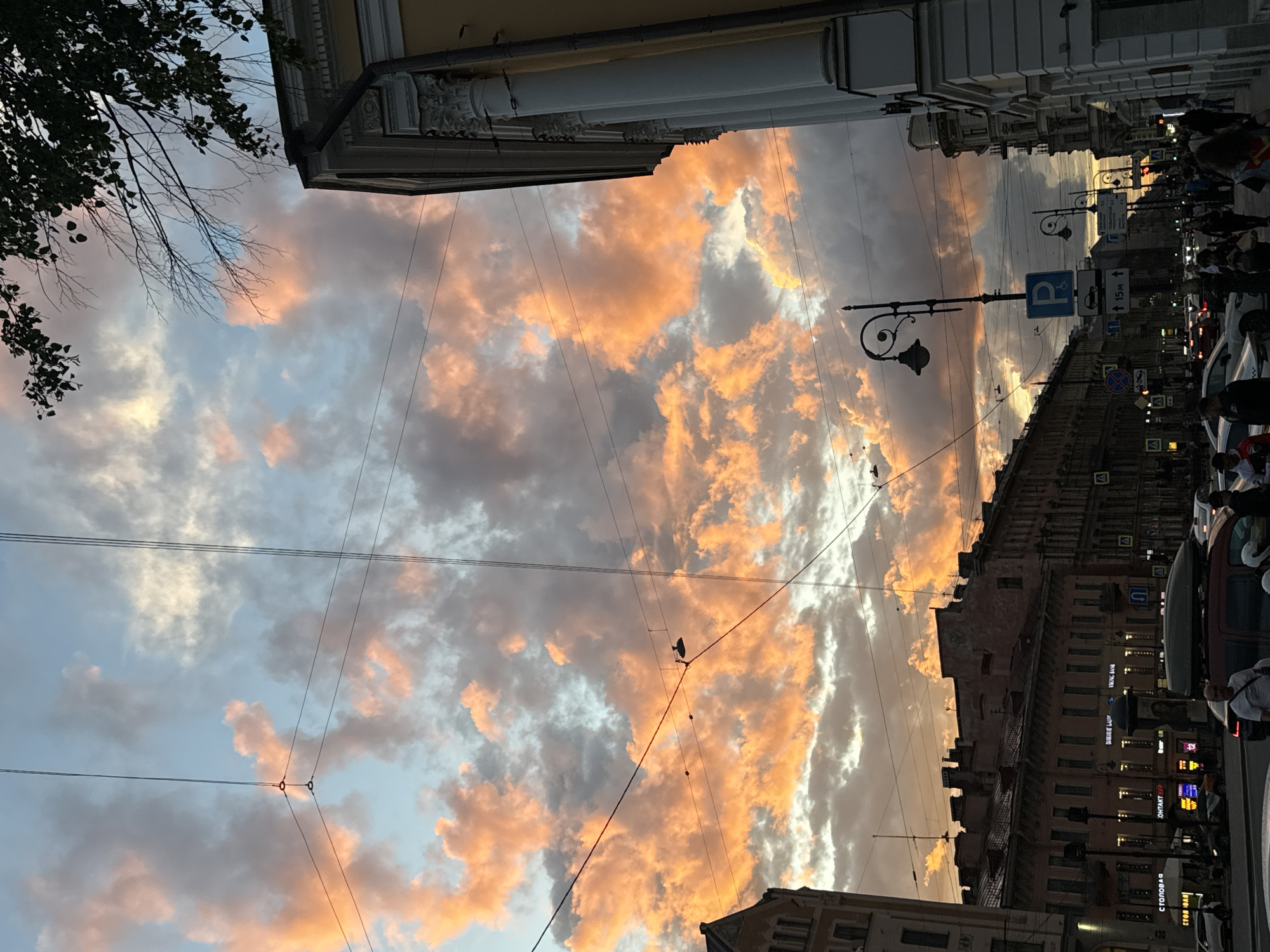 This media file captures a street scene in Saint Petersburg, Russia, during sunset. The sky is filled with dramatic, illuminated clouds, painted in hues of orange, pink, and gray, suggesting a beautiful and perhaps dramatic end to the day.
The perspective is looking up and sideways, giving a slightly disorienting but dynamic view of the urban environment. Overhead, power lines crisscross the frame, adding a layer of texture and a sense of the city's infrastructure.
On the right side of the image, buildings line the street, their facades showing the architectural style typical of Saint Petersburg. Some buildings are illuminated, with lights visible from windows, suggesting that life is continuing inside as the day ends. A street lamp with a decorative design is also visible, a common feature in older European cities.
Further down the street, signs and traffic lights indicate urban activity. A blue parking sign with a P and an electric car symbol is clearly visible. Several cars are parked or in motion, and pedestrians are walking along the sidewalk, creating a sense of a bustling city at dusk. The overall mood conveyed is one of urban beauty, enhanced by the striking natural spectacle of the sunset. The presence of people and vehicles indicates that this is likely a public street or thoroughfare.