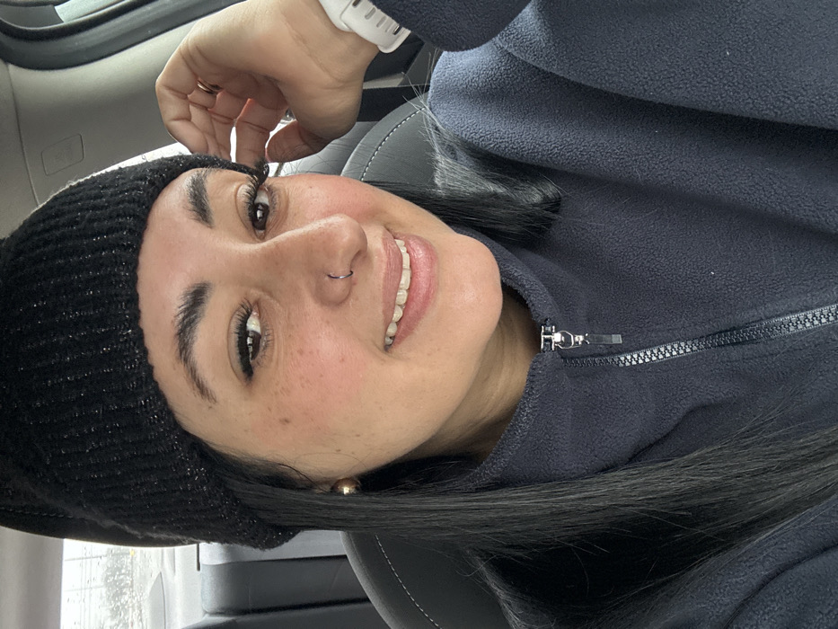 A smiling woman, wearing a black beanie and dark fleece jacket, is indoors. She is fair-skinned with dark hair and eyebrows, wearing a nose ring. The background features a car interior, with a grey leather seat. She is wearing a white smartwatch.