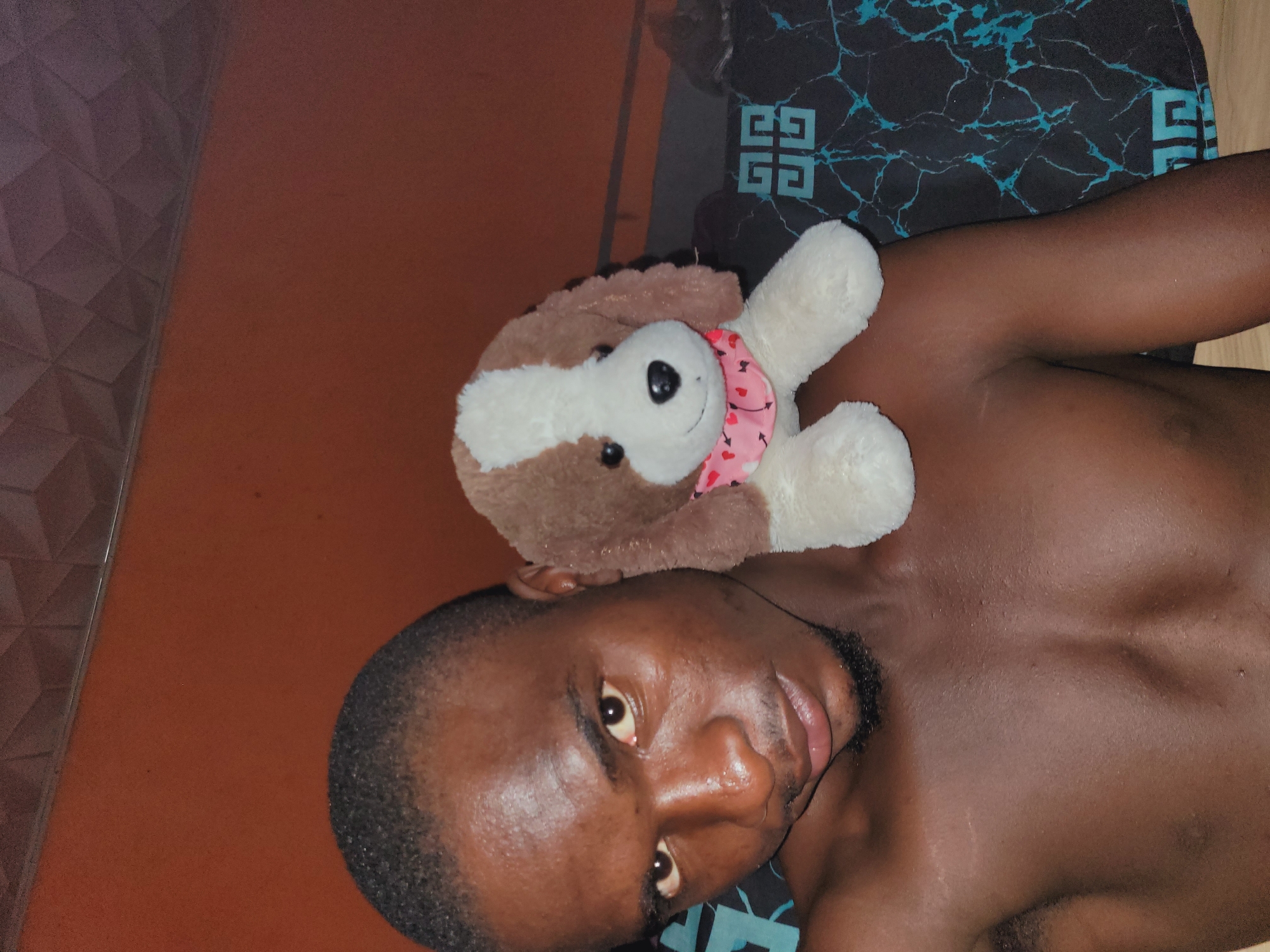 A young Black male is shown from the chest up, with a stuffed dog resting on his left shoulder. He is looking directly at the camera with a slight smile. His skin is dark and appears moist. He has short, dark hair and a goatee. The stuffed dog is brown and white with a pink bandana around its neck. The background is a dark orange wall with a textured ceiling visible at the top. A patterned fabric is visible behind the man and the dog. The scene appears to be an indoor setting, possibly a bedroom. The location in Bukuru, Nigeria, is not discernible from the image itself.