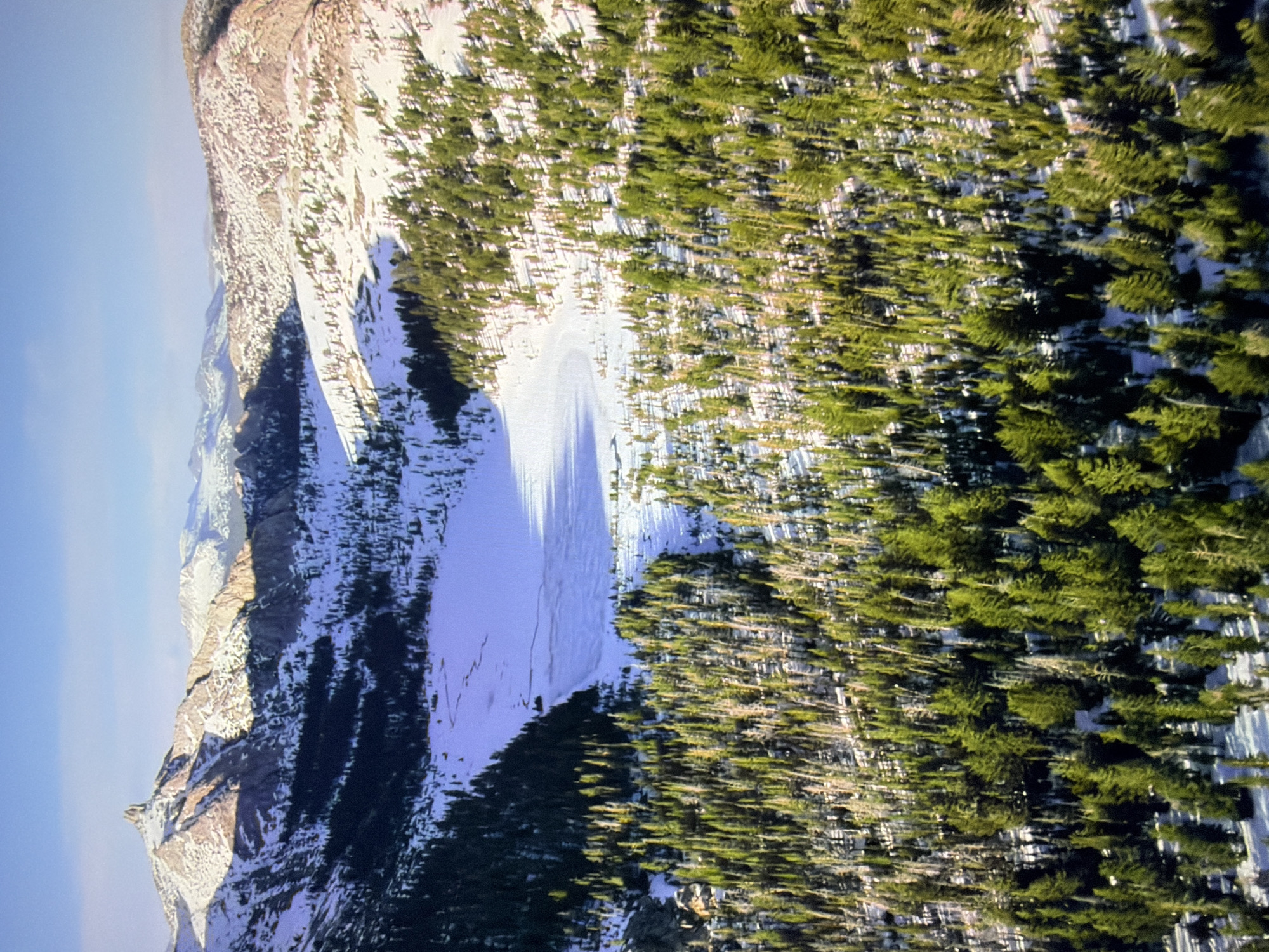 This is a high-angle aerial photograph of a mountainous, snow-covered landscape. In the foreground, a dense forest of evergreen trees dominates the scene, with some trees appearing taller and more established than others. The ground beneath the trees is partially covered in snow, and shadows from the trees are cast across the snow.
In the mid-ground, a large, frozen body of water, likely a lake, is visible. It is predominantly white, indicating snow cover, with some patches of darker ice showing through. A winding path or stream is faintly visible on the surface of the ice. The edges of the lake are bordered by more evergreen trees and snow-covered terrain.
The background features a series of majestic, snow-capped mountains. The prominent peak on the left has a sharp, distinctive summit that catches the sunlight. The mountains are rugged and appear to be a mixture of rock and snow. The sky above is clear and a pale blue, suggesting a bright, cold day.
The image depicts a remote, natural environment, likely a wilderness area in a mountainous region during winter. There are no people, man-made structures, or any indication of urban environments, and therefore no connection to the city of Kyiv, Ukraine.
