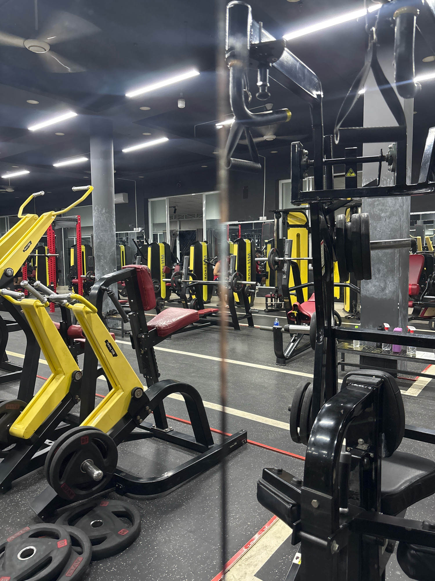 This is an indoor image of a gym located in Nha Trang, Vietnam. The gym is equipped with various weightlifting machines and equipment, including a squat rack with weight plates, a bench press, and several yellow-framed weight machines. The space is well-lit by fluorescent lights on the ceiling, and ceiling fans are also visible. The floor is dark-colored with white and red markings. In the background, mirrors line one wall, reflecting more of the gym equipment. There are no people clearly visible in the foreground, but a few individuals can be seen in the reflected background. The overall impression is of a functional and modern fitness facility.