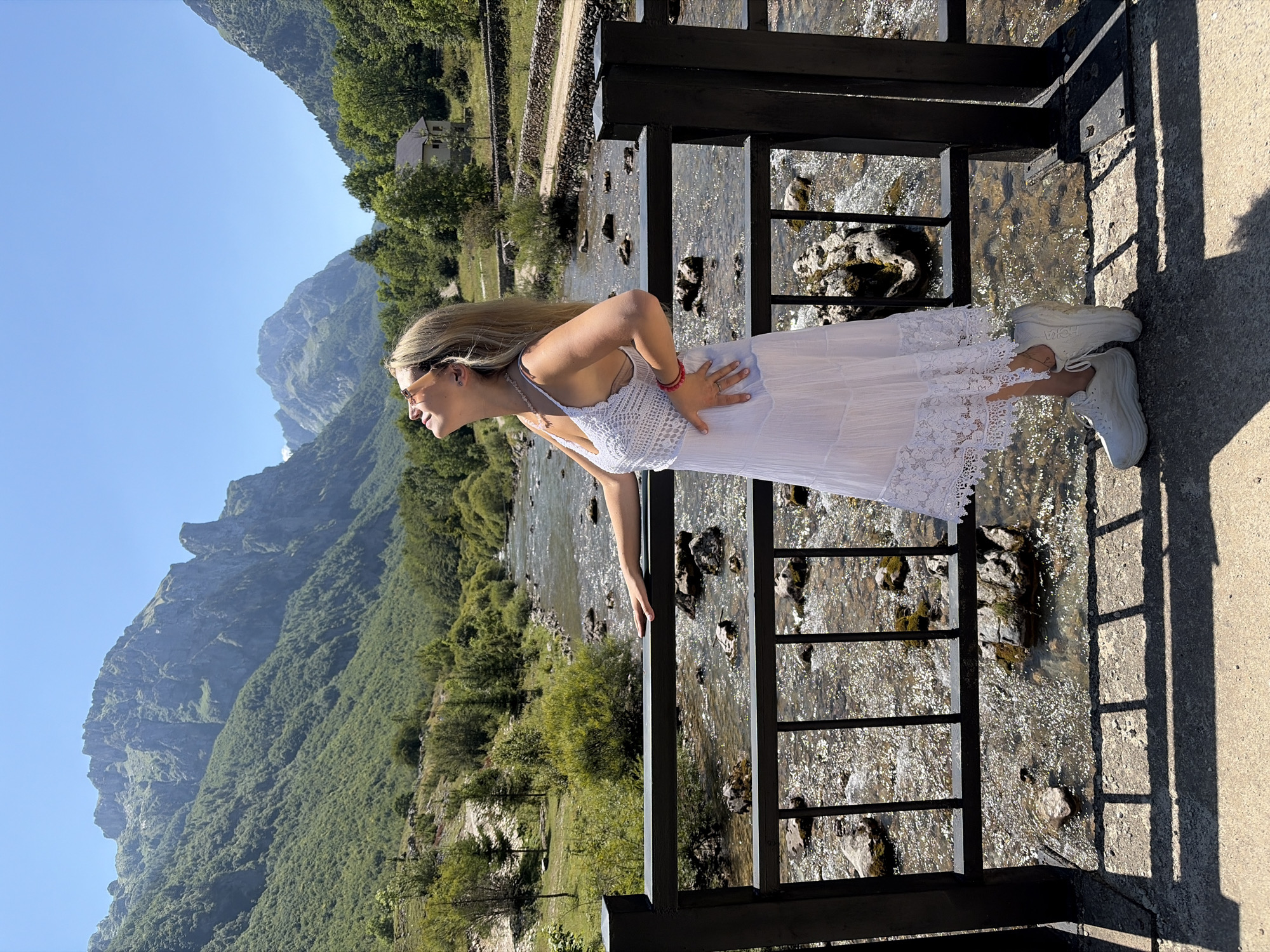 A young woman with blonde hair stands on a bridge overlooking a river in Vusanje, Montenegro. She is wearing a white sundress and white sneakers, with her head tilted back and eyes closed as if enjoying the sun. The river flows over rocks, and the background features lush green mountains and a clear blue sky. A small house is visible in the distance.