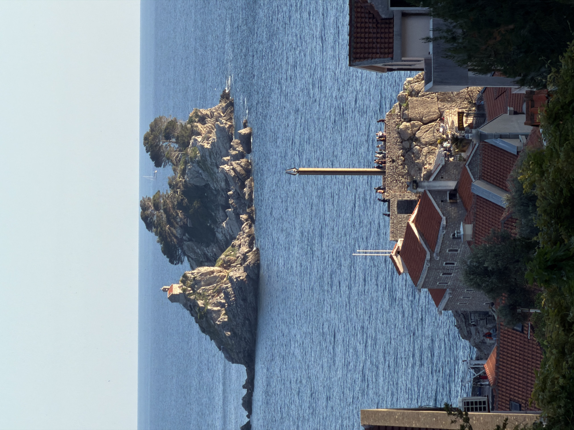 The image shows a coastal scene in Petrovac na Moru, Montenegro. Two rocky islands rise from the Adriatic Sea. The island on the left features lush vegetation and a small structure at its base. To the right, connected to the mainland by a narrow isthmus, is a cluster of buildings with red-tiled roofs, some with antennas, interspersed with trees. People can be seen on the rocks along the isthmus. The sea stretches between the islands and toward the horizon under a bright sky. The image is vertically oriented.