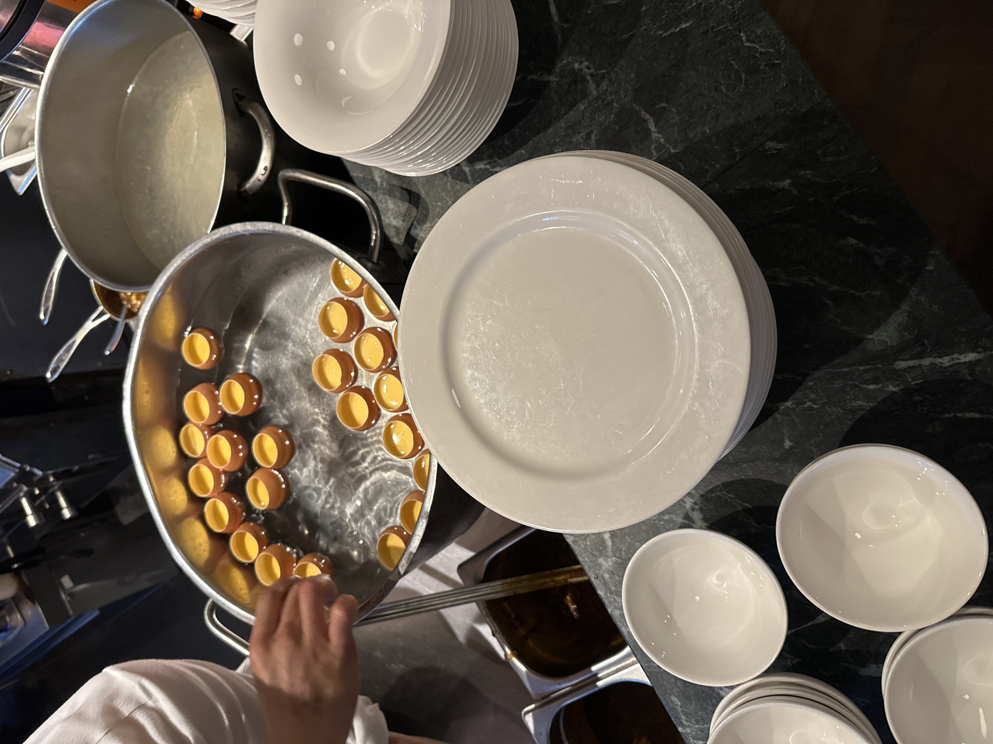 The image depicts a kitchen scene, with a focus on food preparation. A chef in a white uniform is seen using tongs to handle small, filled ramekins in a large pot of water. Adjacent to the pot, there are stacks of white plates and bowls, suggesting an assembly line-like setup. Two containers with dark, liquid contents are visible as well. The kitchen's countertop has a dark, marble-like appearance. There is no information available within the image itself regarding the location of this kitchen within London, United Kingdom, nor the broader external environment.