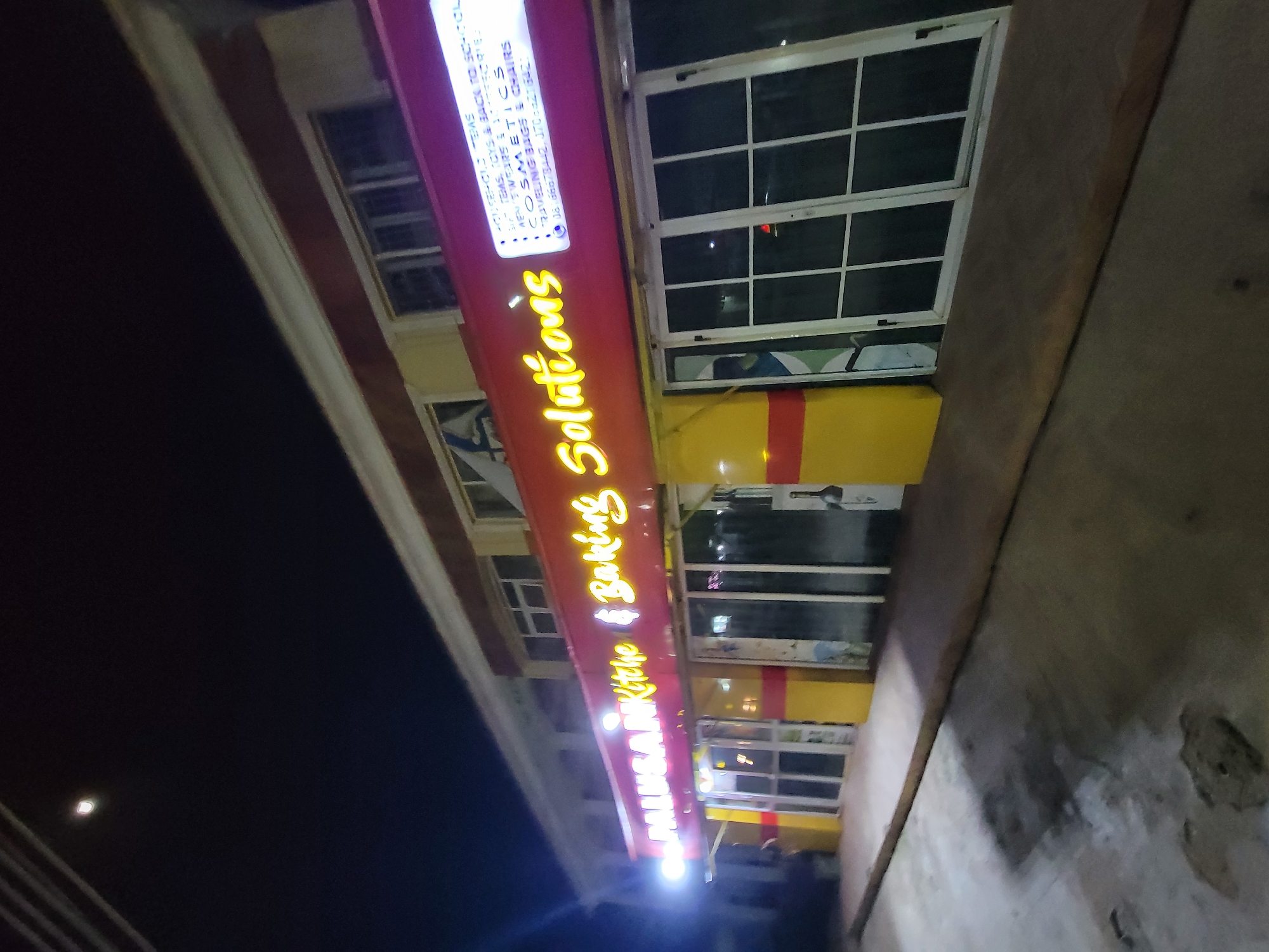 The image captures an outdoor view of the "MALUSAR Kithe & Baking Solutions" store at night. The storefront features a red sign with bright yellow lettering. The sign also lists various product categories, including "HOUSEHOLD TEMS", "STEMS, TOYS & BACK TO SCO-COL", "MENSWEAR SSORIES", "COSMETICS", and "RAVELING BAGS & CHAIRS". The store's windows are visible behind the sign, with a yellow and red color scheme underneath. The location is in an unidentified city, Unknown.