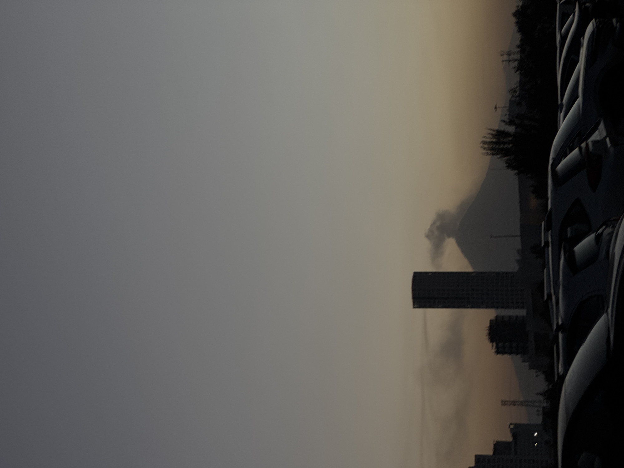 The image presents an urban landscape bathed in the dim light of what appears to be dusk or dawn, characterized by a pervasive haze. The sky transitions from a soft, yellowish-orange tint near the horizon to a muted grey overhead.
In the right foreground, several dark-colored vehicles, likely SUVs or vans, are parked, their forms reduced to strong silhouettes. Reflections on their surfaces reveal minimal detail.
The midground features prominent architectural elements. A tall, dark rectangular skyscraper stands centrally, its windows indistinct against the hazy sky. Below and to its right, other smaller, blocky buildings and what looks like a construction crane are partially visible, also in silhouette.
The background reveals a large mountain range, its peaks partially obscured by the widespread haze. A distinctive plume of white-grey smoke or cloud rises from behind or near one of the mountains on the left, contributing to the atmospheric conditions. Silhouetted trees dot the landscape along ridges, and a faint power line tower can be seen on a distant hill.
The scene is devoid of human activity or visible text. The combination of urban structures, natural mountain scenery, and the rising smoke creates a dramatic and somewhat somber atmosphere, consistent with views of a city like Puebla, Mexico, potentially under the influence of volcanic activity from nearby Popocatépetl or environmental factors.