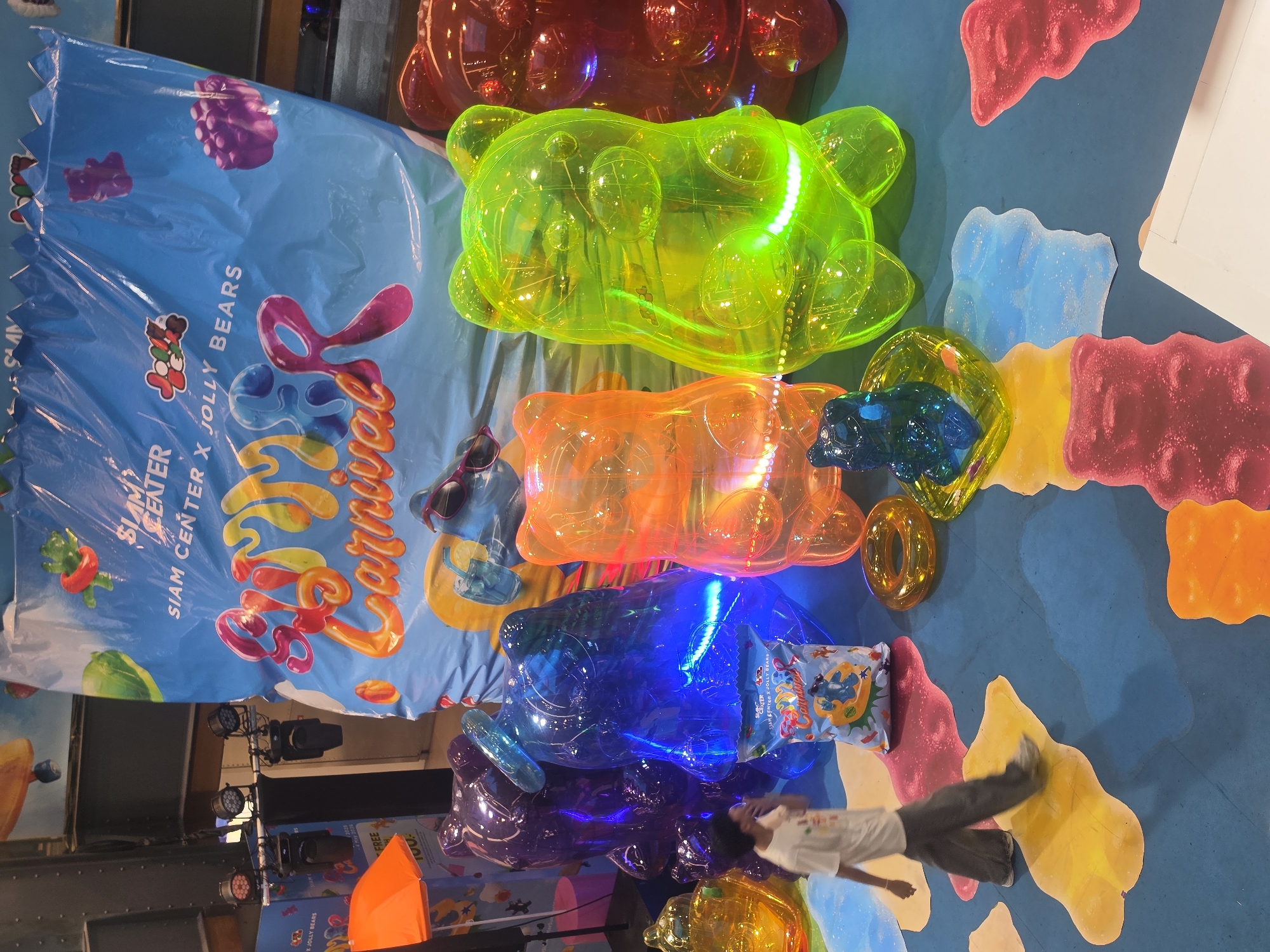 The image depicts an vibrant indoor exhibition or event, likely a "Carnival" collaboration between "Siam Center" and "Jolly Bears" in Bangkok, Thailand. The central subjects are several large, translucent gummy bear sculptures, each internally lit, arranged along a blue floor.
From top to bottom, the visible gummy bear sculptures are red, glowing neon green, bright orange, deep blue, and purple. Their transparent bodies and internal illumination create a luminous, candy-like effect. A smaller, dark blue gummy bear figure and a gold ring-shaped object are also visible among the larger sculptures.
A large blue banner hangs vertically on the left, serving as prominent signage for the event. It clearly displays the text "SIAM CENTER X JOLLY BEARS Carnival" in a colorful, stylized font, along with cartoonish fruit and gummy bear graphics. The banner also notes "FREE" and dates "20 - 28 MAY," indicating the event period. In the background, stage lighting fixtures are visible, suggesting a professional exhibition setup.
The blue floor is decorated with various flat, brightly colored shapes resembling melted gummy candies in shades of red, yellow, light blue, and pink, complementing the gummy bear theme. A person, partially visible in the bottom right, wearing a white t-shirt and dark pants, walks across the floor, providing a sense of scale to the oversized sculptures. A small, rectangular pillow-like object with "Carnival" branding is also present near the blue gummy bear. The overall aesthetic is whimsical, playful, and brightly colored, characteristic of a fun, public-facing event.
