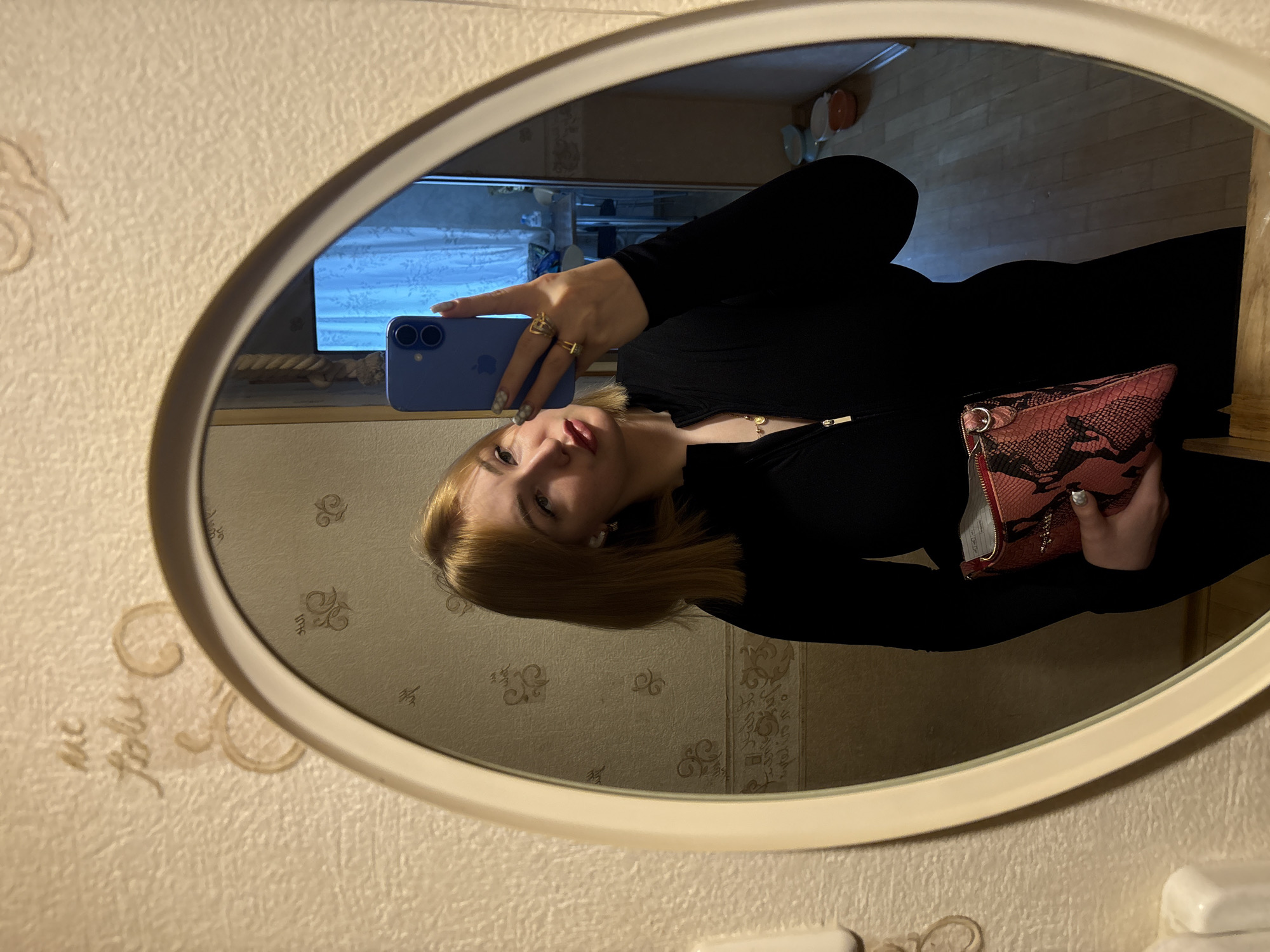 A young woman with blonde hair and red lipstick is taking a selfie in an oval mirror. She is wearing a black zippered top and holding a pink and black snakeskin clutch bag. She has several rings on her fingers. The mirror is on a wall with textured wallpaper featuring a subtle floral pattern. In the reflection of the mirror, a room is visible with wooden flooring, a table, and a window with blue curtains. A rope hangs on the wall in the background of the reflection. The text "me folw" is inscribed on the wall above the mirror.
