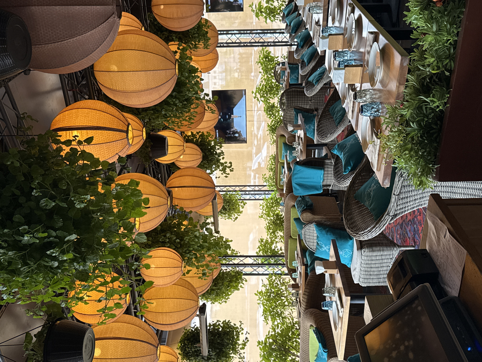 The image shows the interior of a restaurant or cafe, possibly located in Moscow, Russia. An outdoor terrace or patio is seen, with a long table set for a meal. Chairs are arranged on either side of the table, each with a teal pillow. The table is decorated with plates, glassware, and decorative plants.
The ceiling above is covered with decorative lamps and hanging greenery. A TV screen is visible in the background. The atmosphere appears to be upscale and inviting. The presence of plants and decorative lighting suggests a focus on aesthetics and ambiance.