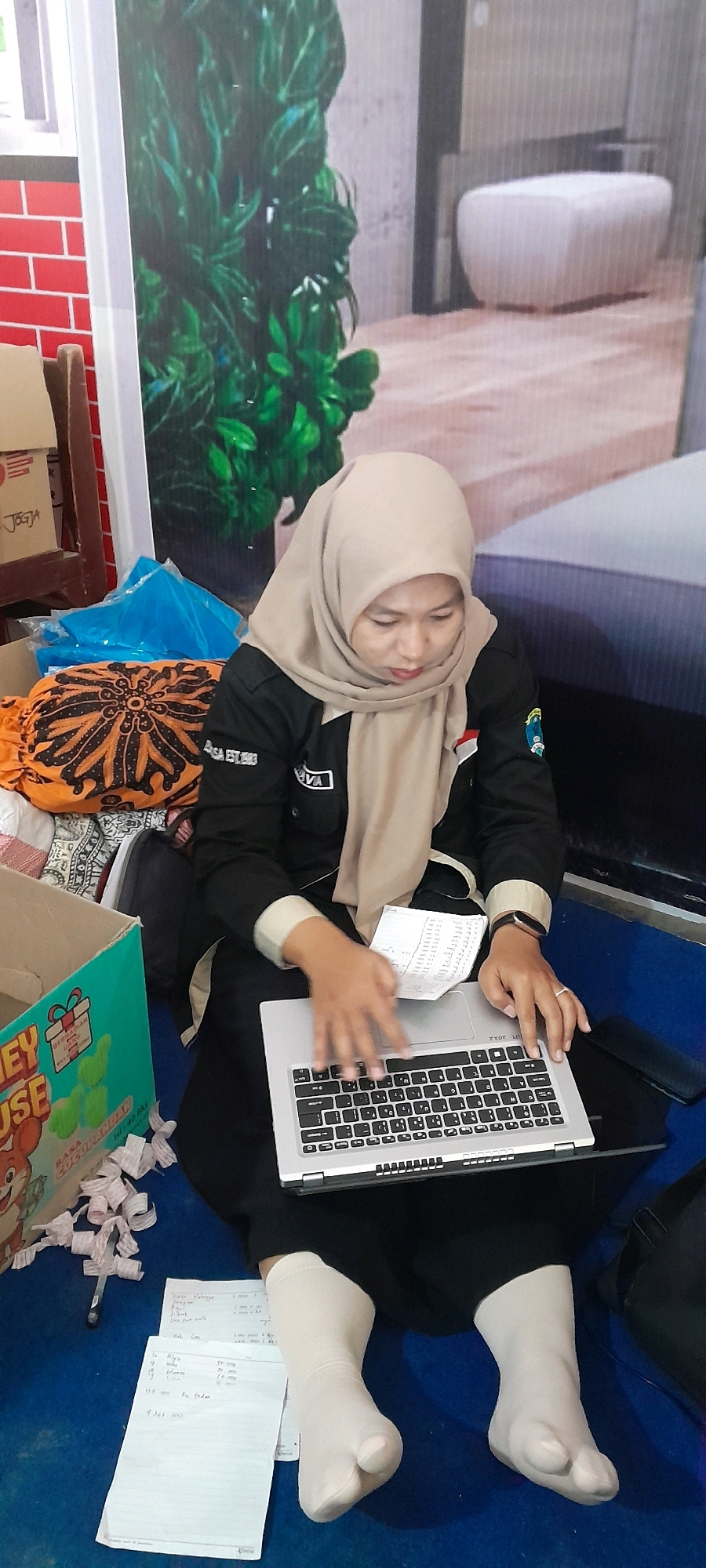 A woman wearing a beige hijab and black uniform with a white collar is seated on a blue floor in what appears to be an indoor space in Tahunan, Indonesia. She is looking down at a silver laptop placed on her lap, with her hands on the keyboard. In front of her, on the floor, are several pieces of white paper with handwritten text. To her left, there is a green cardboard box with a cartoon mouse image and the text "MONEY HOUSE" on it. Behind her, an orange cloth with a black and brown intricate pattern is visible, along with some blue plastic sheeting. The background features a large wall graphic depicting lush green plants and part of a modern interior with a white ottoman. The lighting is dim, creating shadows on the floor.