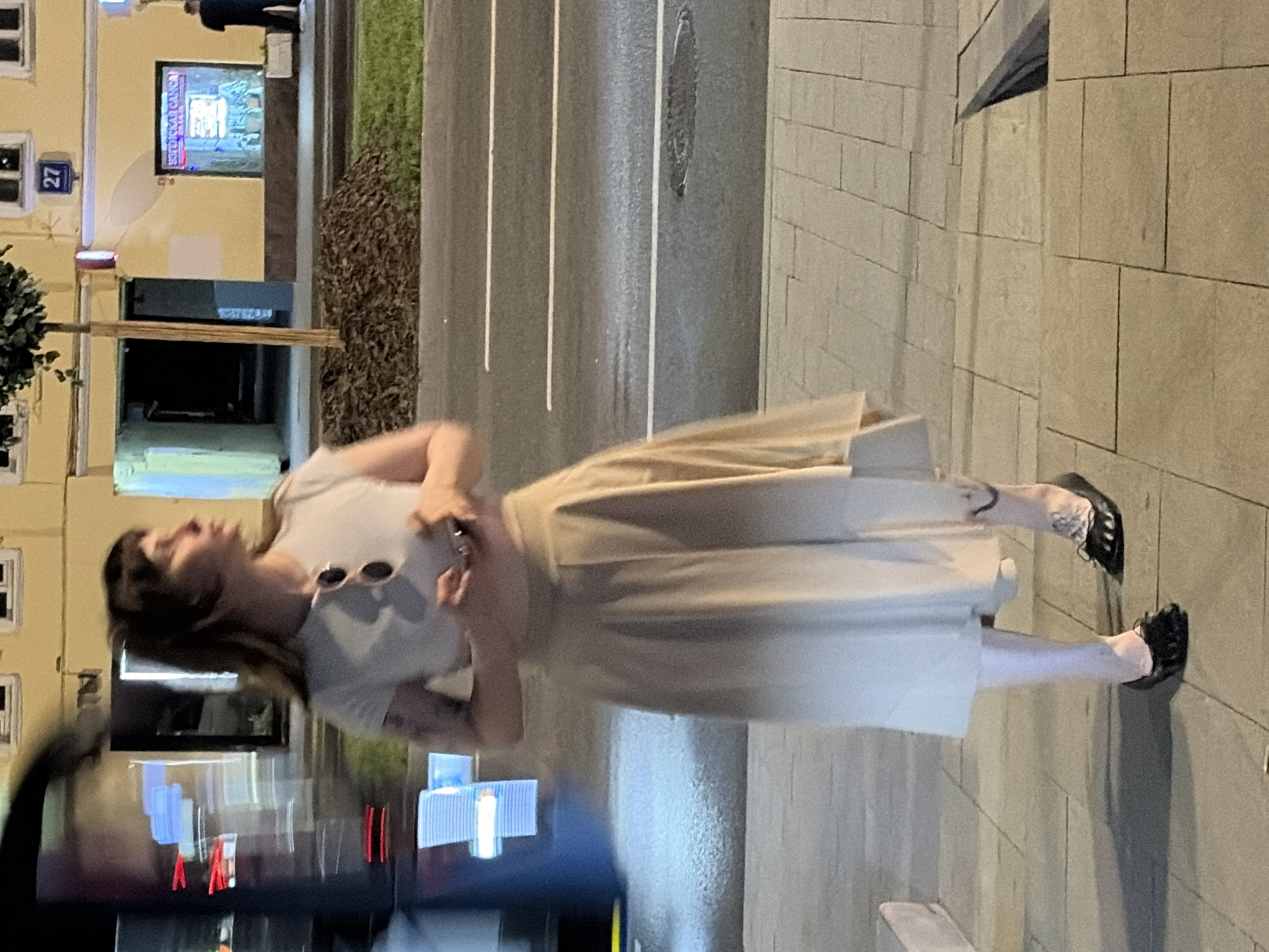 A young woman is standing on a sidewalk at night in front of a blurred moving vehicle. She has dark hair, is wearing a light-colored crop top, a long, flowing skirt in a gradient of beige to white, and black flat shoes. Sunglasses are pushed up on her head. She is holding a mobile phone in her hands. Her left leg has a tattoo visible. The sidewalk is paved with light-colored rectangular tiles. Across the street, there are buildings with yellow facades, some with illuminated windows. A street sign numbered "27" is visible on one of the buildings. A small tree with dark mulch around its base is planted on the sidewalk. The street in front of the sidewalk has visible white lane markings and appears wet, reflecting some light. The scene is likely in a city environment, consistent with Moscow, Russia given the context.