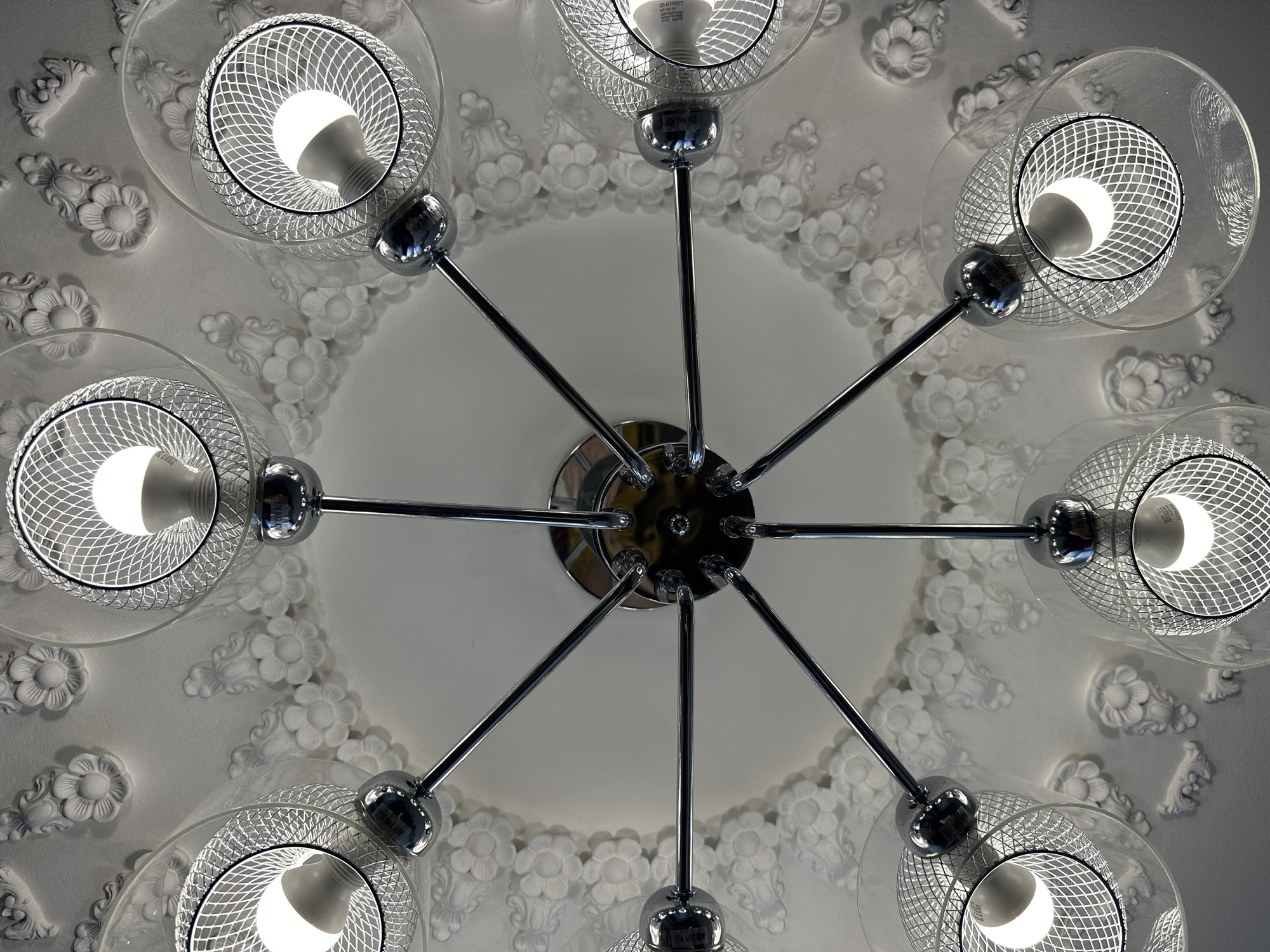 The image shows a chandelier with eight arms extending from a central fixture. Each arm holds a light fixture with a clear glass shade and a silver mesh interior. Inside each shade, a bright LED bulb is visible. The chandelier is mounted on a white ceiling adorned with intricate floral plasterwork. The perspective is looking directly up at the chandelier from below. The location in the city of Yessentuki, Russia, is not discernible from the image itself. There are no people or actions depicted.