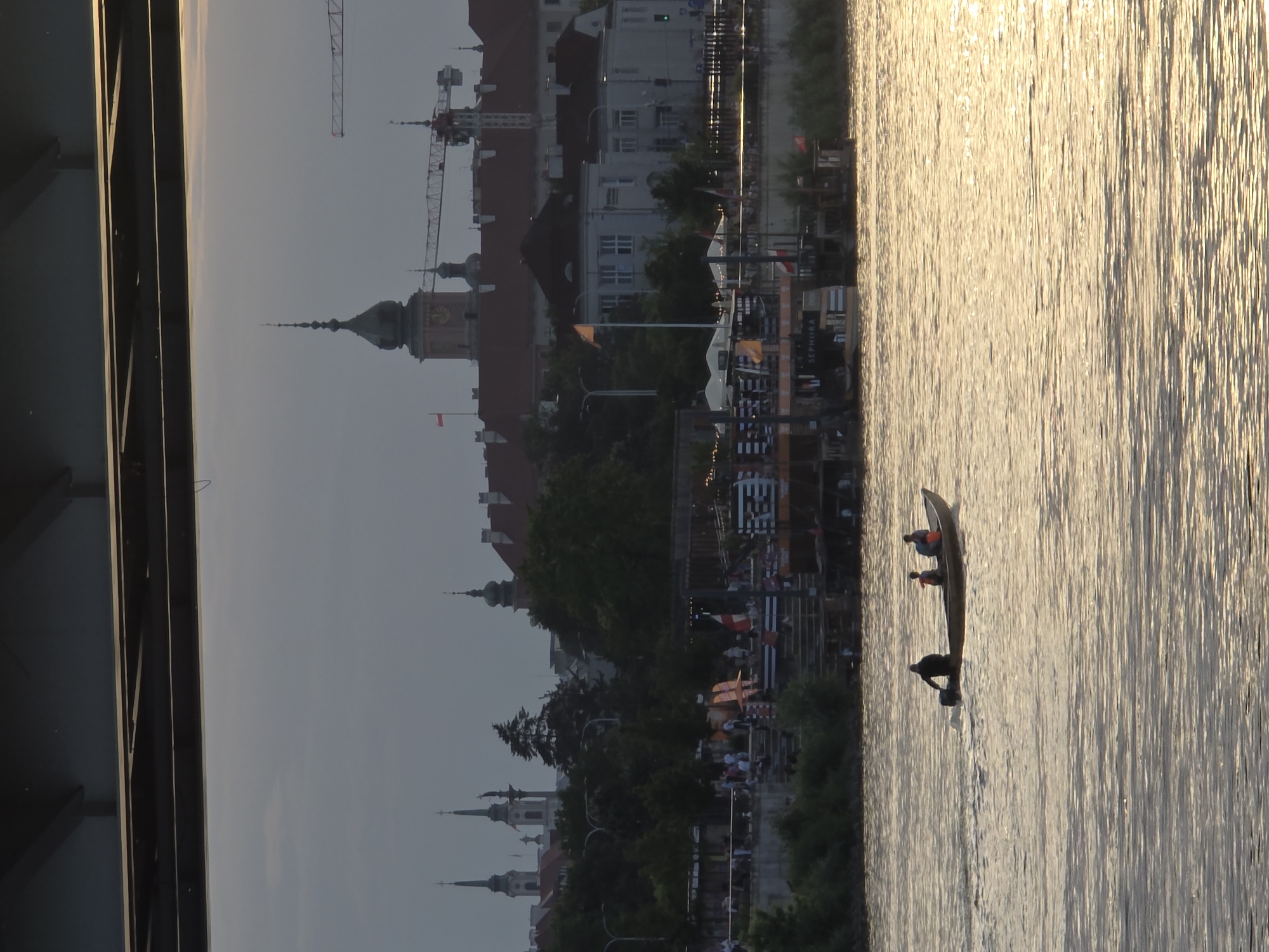 The image depicts a waterfront scene in Warsaw, Poland, during what appears to be sunset. The sun's golden light reflects off the rippling surface of a wide river, creating a shimmering effect. In the foreground, a narrow boat with two people aboard navigates the water. One person is seated, while the other is partially visible, perhaps steering or paddling.
Beyond the river, the cityscape of Warsaw is visible. Buildings with traditional architecture, including domed structures and spires, rise above lush green trees. Cranes, indicative of ongoing construction or development, are also present. Along the riverbank, there are signs of activity, with what appears to be an outdoor market or event space, featuring stalls and people gathered. The presence of flags suggests a festive atmosphere.
The time of day is clearly indicated by the low angle of the sun and the warm, diffused light, casting a soft glow over the scene. The sky is a pale gray, transitioning towards dusk. The overall mood is peaceful and serene, with a touch of urban vitality. The image is captured from a slightly elevated perspective, possibly from a bridge or a building overlooking the river. No specific text is clearly legible in the image, although there may be some signage on the structures in the background that is too small to decipher.