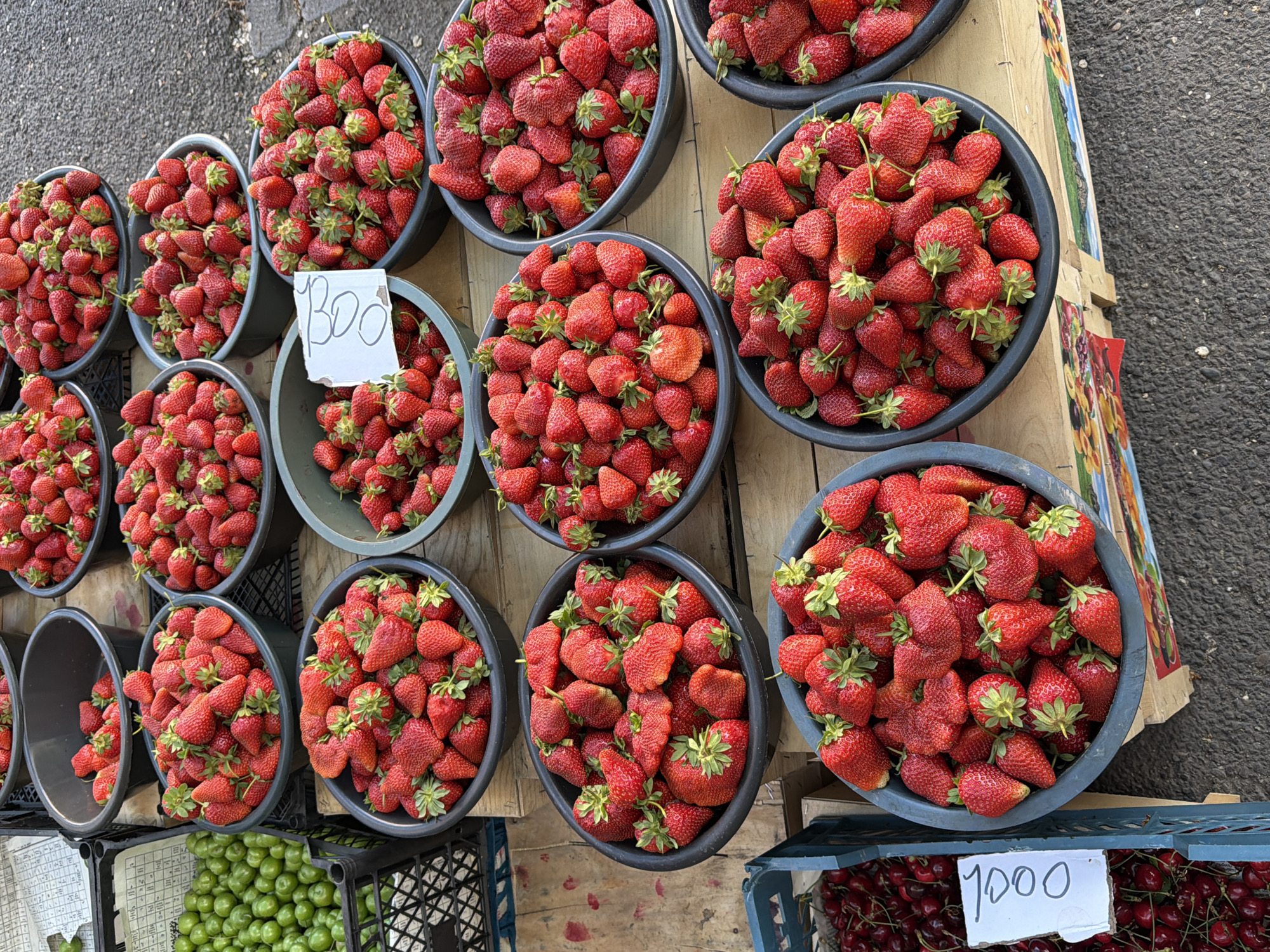 The image depicts a fruit stand in Yerevan, Armenia. Eleven bowls of strawberries are arranged on a wooden structure, with price signs indicating "800" and "1000." Crates containing green plums and cherries are visible beneath the strawberries. The background includes a paved surface and parts of the vendor's setup.