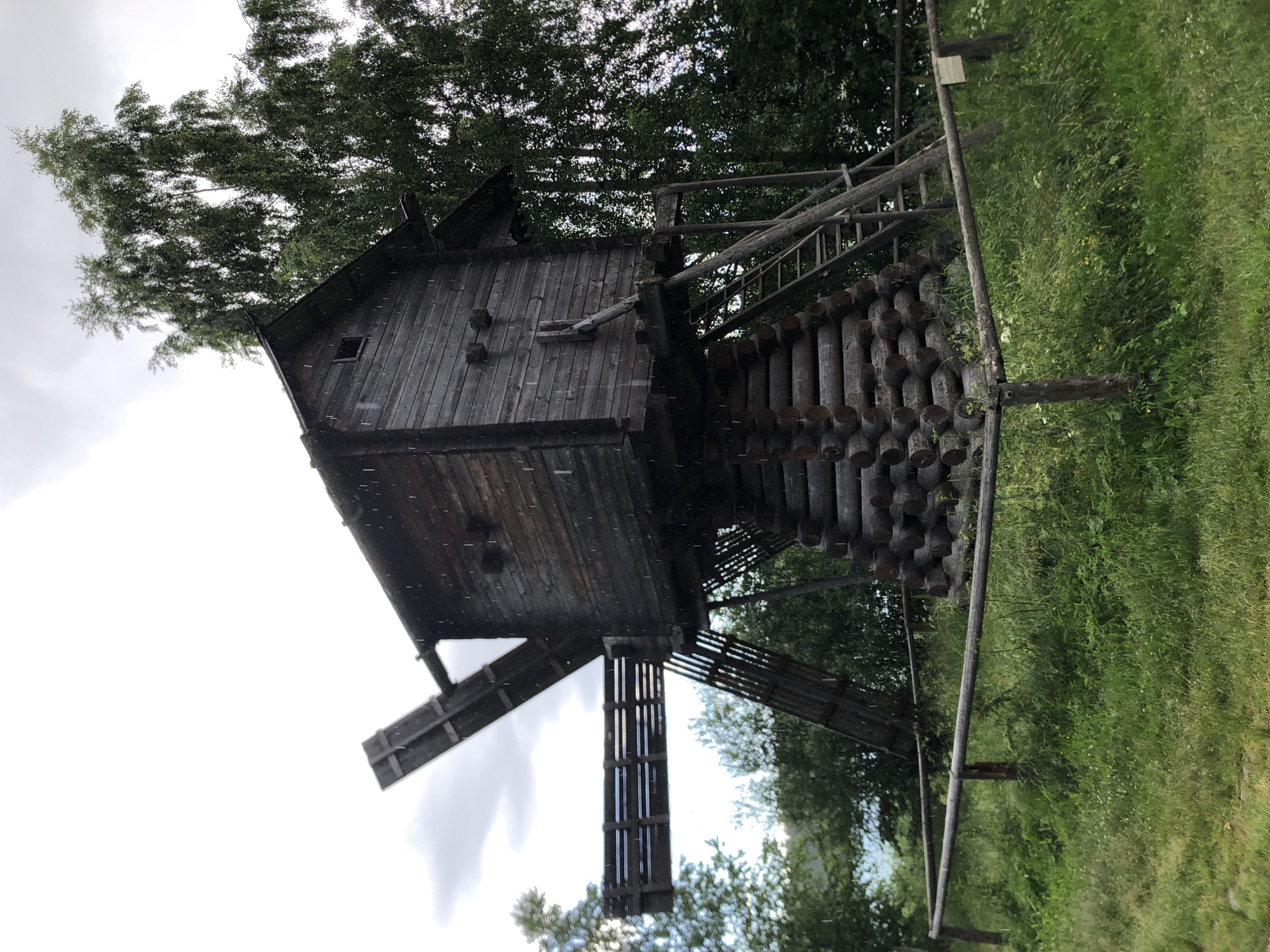 The image depicts an outdoor scene in Verkhniye Mandrogi, Russia, featuring a rustic, weathered wooden structure resembling a windmill. The structure is made of dark, aged wood and appears to be elevated on a sturdy base constructed from vertically stacked logs. A wooden staircase leads up to the main body of the windmill, which has a small, dark-framed window and a peaked roof. A wooden beam extends outwards from the upper part of the structure. The windmill is situated amidst lush green grass and foliage, with trees visible in the background and overhead. The sky is overcast, suggesting a cloudy day. There are visible specks throughout the image, which could be raindrops or atmospheric particles. The overall impression is one of a historical or traditional building set in a natural environment. No people are visible, and no text is present in the image. The scene conveys a sense of rural tranquility and historical preservation.