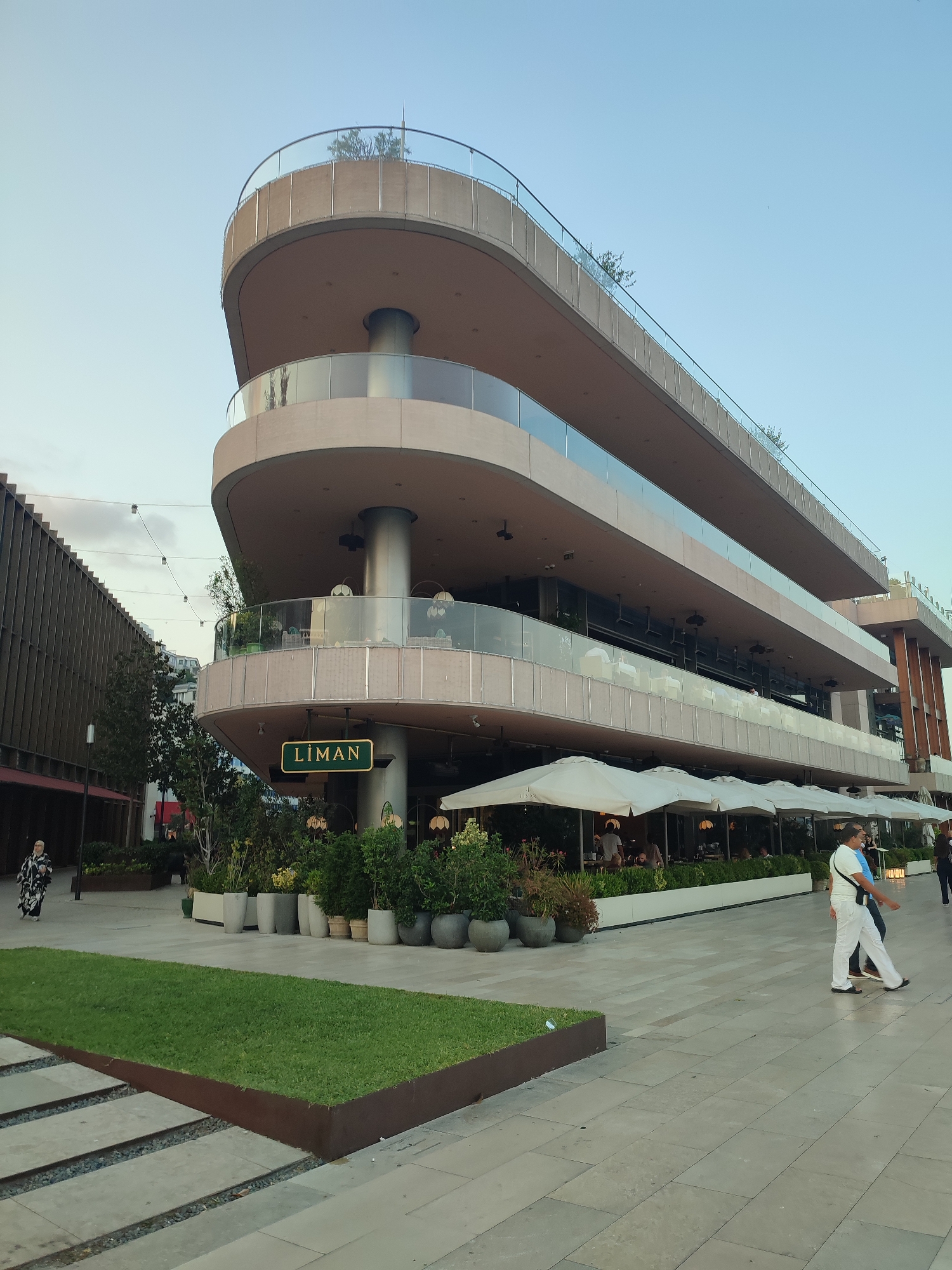 The image shows a multi-story building with a curved facade and balconies, located in the city of Beyoğlu, Turkey. The building appears to house a restaurant or cafe, indicated by the sign "LİMAN" and the presence of outdoor seating with umbrellas. Several people are walking on the paved walkway in front of the building. The area also features green spaces with grass and potted plants. In the background, another building with a slatted facade is visible. The sky is clear and blue.