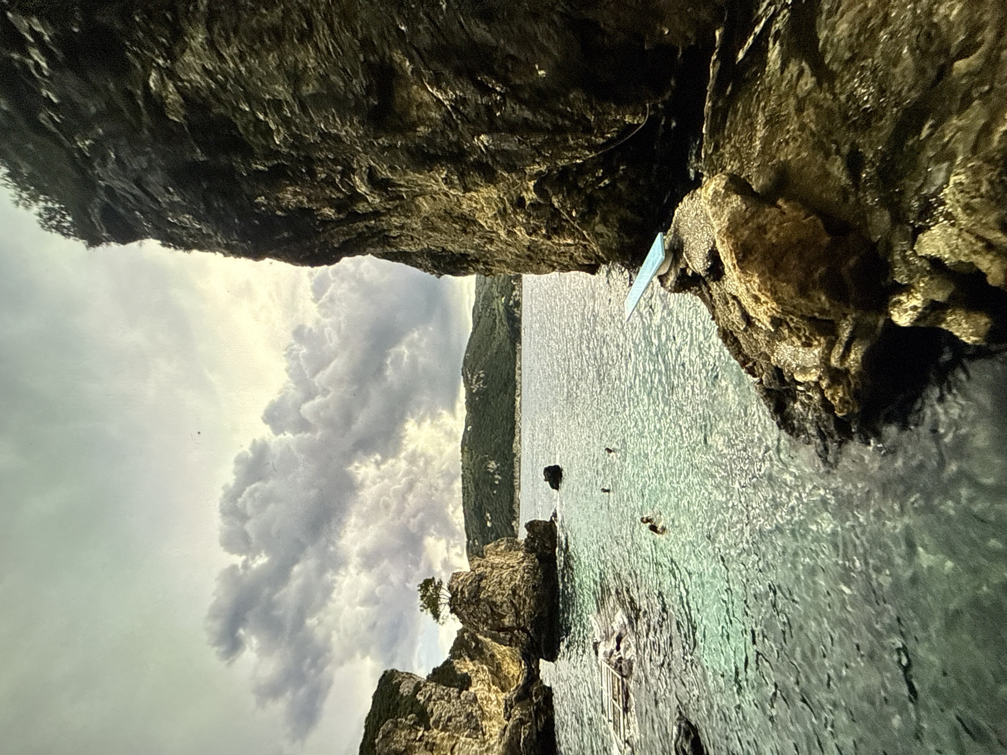 The image presents a view of a rocky coastline with an overcast sky. The frame is dominated by the rugged cliffs on either side. The sea fills the central area of the image, with scattered rocks emerging from the water near the shoreline. Several people appear to be swimming in the water. A diving board is visible extending from one of the cliffs. The location is not Sofia, Bulgaria.