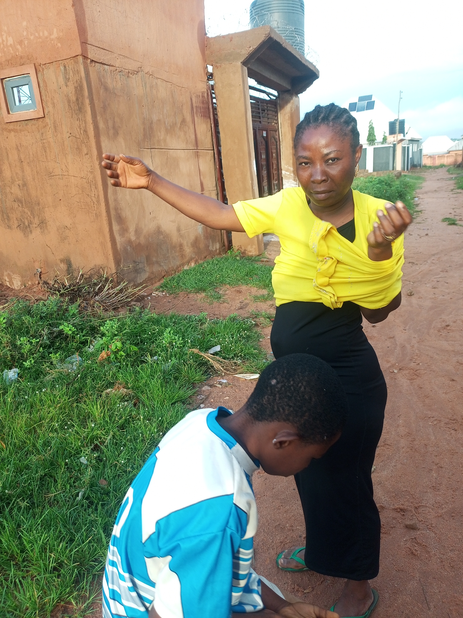 A woman, facing the camera and wearing a bright yellow shirt knotted at her waist and a long black skirt, stands on a dirt road in Tempure, Nigeria. Her left arm is extended towards a building on her left, her hand open, as if gesturing. Her right hand is raised and clenched. She has braided hair. A young child, seen from the back, is positioned in front of her, looking down. The child wears a blue and white shirt with horizontal stripes on the sleeves. Green grass grows on the left side of the road, with some debris visible. In the background, a brown building with a small window and a water tank on its roof is partially visible, along with other structures and vegetation. The sky is overcast.
