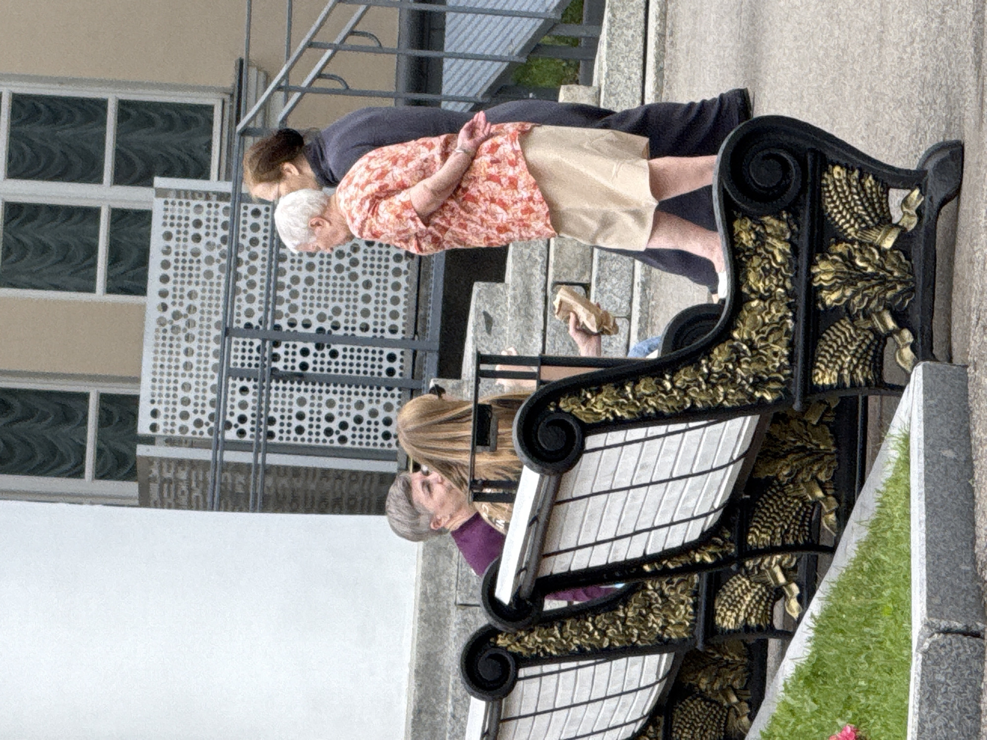 A group of people are gathered on an outdoor paved area with benches and a staircase. In the foreground, ornate black and gold benches are visible, with a patch of green grass to the left. Two individuals are seated on the bench in the foreground, partially obscured. Behind them, to the right, two women are standing. One, with short gray hair, faces towards the viewer's left. The other, with long brown hair, faces away from the viewer. Another woman with white hair and a floral print shirt and a beige skirt is walking away from the viewer, with her hands clasped behind her back. A person's hand holding a brown paper bag is visible near the standing women. The background shows a building with windows and a decorative metal grate. The scene appears to be a public space in Moscow, Russia, possibly a park or the grounds of a cultural institution.