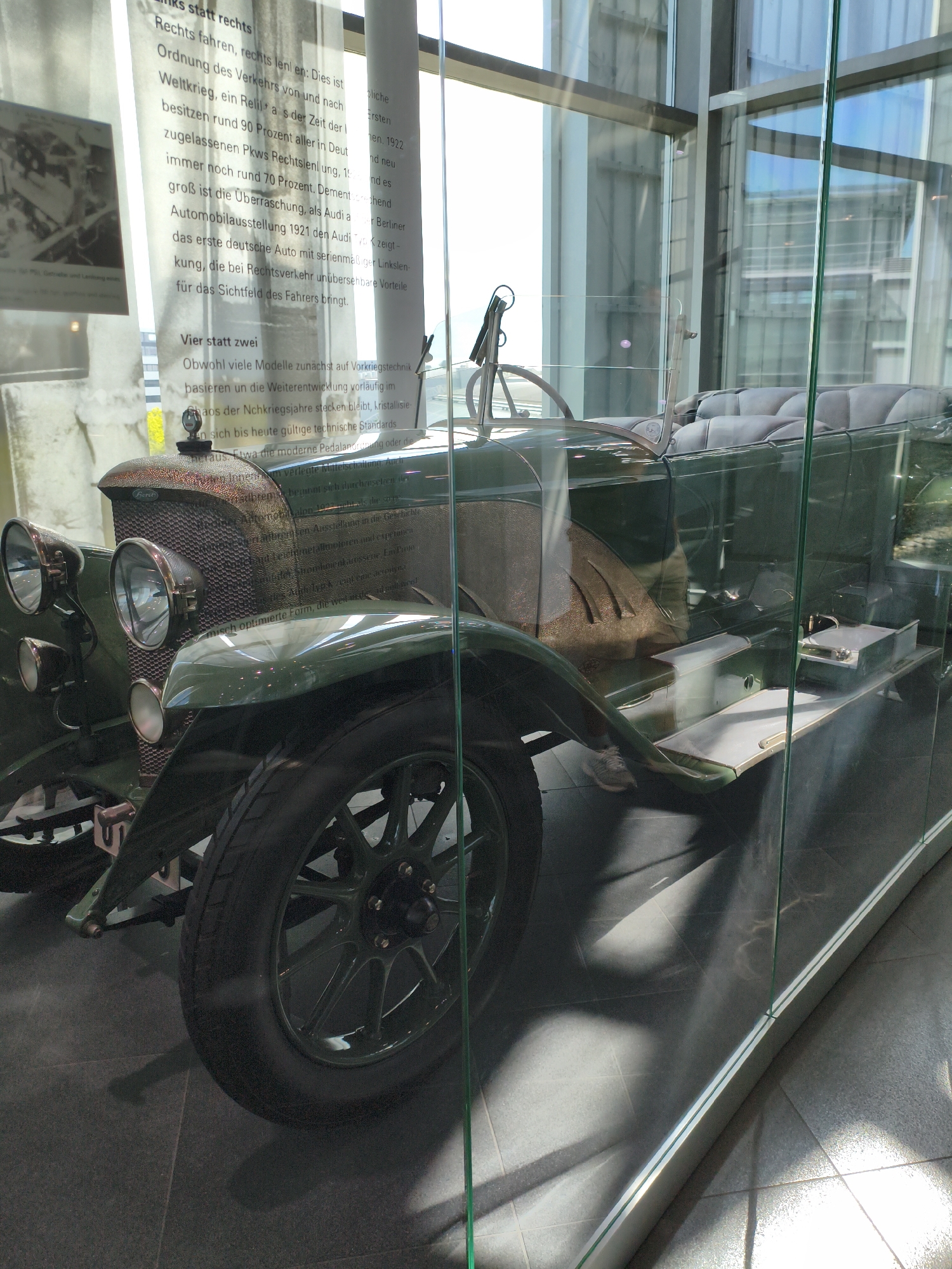 An antique green vehicle is displayed behind glass in a museum. The text to the left of the vehicle provides historical context about early automobiles and traffic regulations in Germany. The vehicle itself is a four-wheeled automobile with a convertible top and a spoked steering wheel. It has prominent headlights and a visible engine grille. The surrounding environment appears to be a modern museum with glass walls and tiled floors. The lighting suggests it is daytime, with natural light filtering in. The text describes the transition from right-hand to left-hand driving, the impact of World War I on the automotive industry, and the innovations introduced by early Audi models. The text also discusses the evolution of vehicle design from post-war technology to more modern standards.