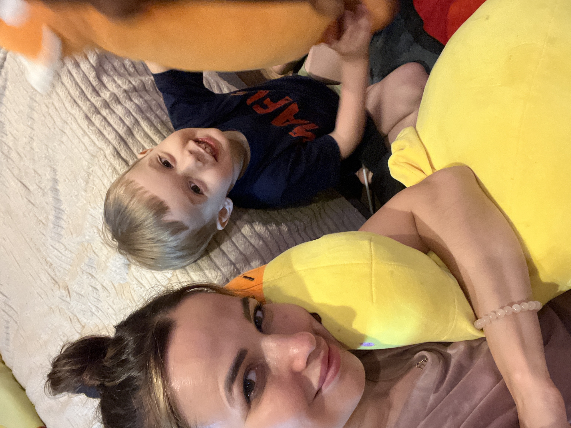 An overhead, close-up shot shows a young boy and a woman, likely his mother, lying on a textured, beige bedspread, holding large stuffed animals. The woman, on the left, has her hair in a bun and is smiling at the camera, holding a large yellow duck plushie. The boy, on the right, is wearing a navy blue t-shirt and is smiling widely. He is holding a large, orange plushie. The scene suggests a casual, indoor setting, possibly a bedroom.