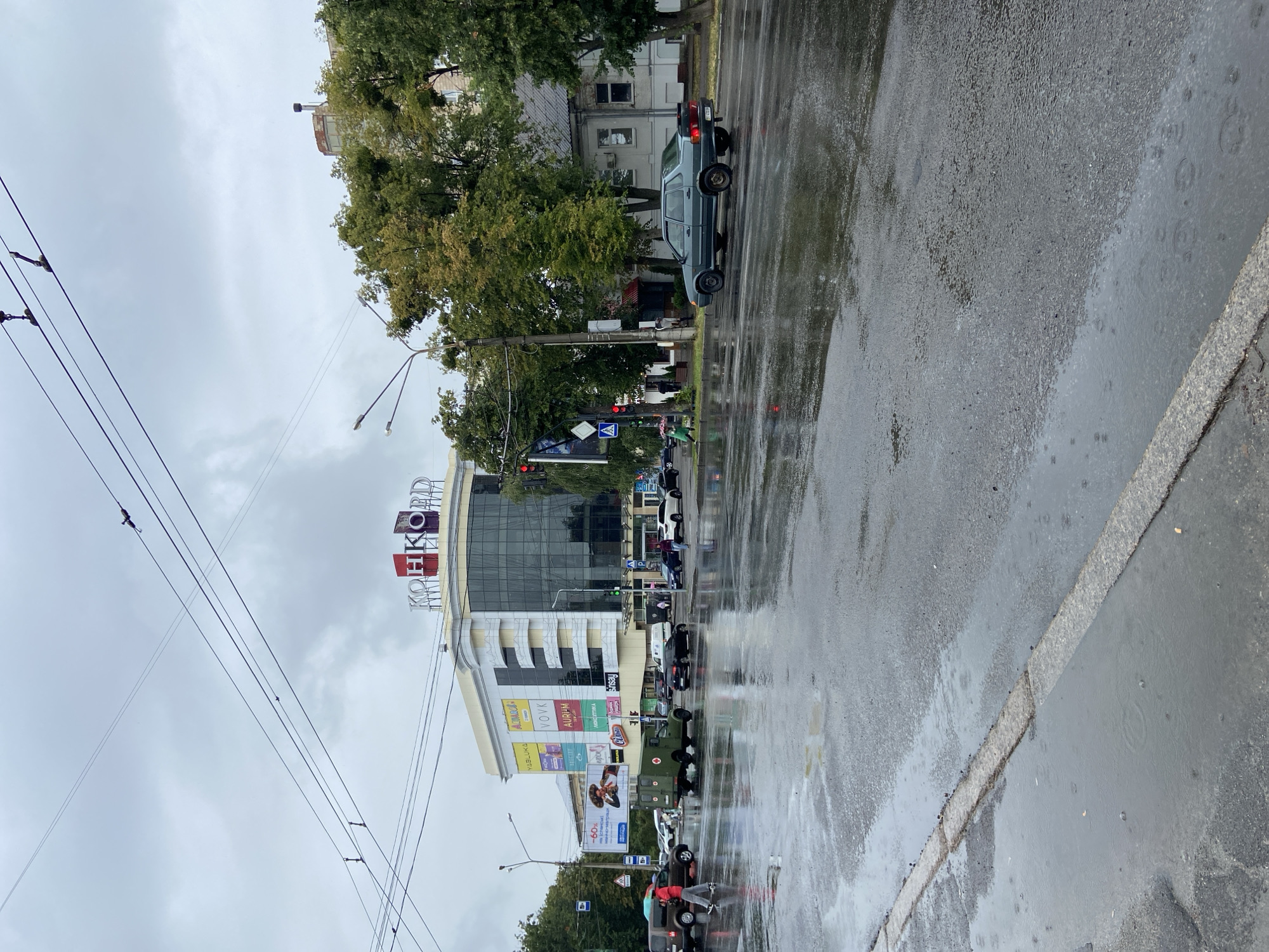 The image shows a street scene in Nyzhni Mlyny, Ukraine on a rainy day. A large shopping center with the sign "КОНКОРД" (Konkord) is visible in the background. Various cars, including a dark green military-style vehicle, are on the wet, reflective asphalt street. Pedestrians are also present, some carrying umbrellas. The sky is overcast, and there are power lines overhead. The environment is urban, with buildings and trees lining the street. Puddles of water are on the road, indicating recent rainfall.