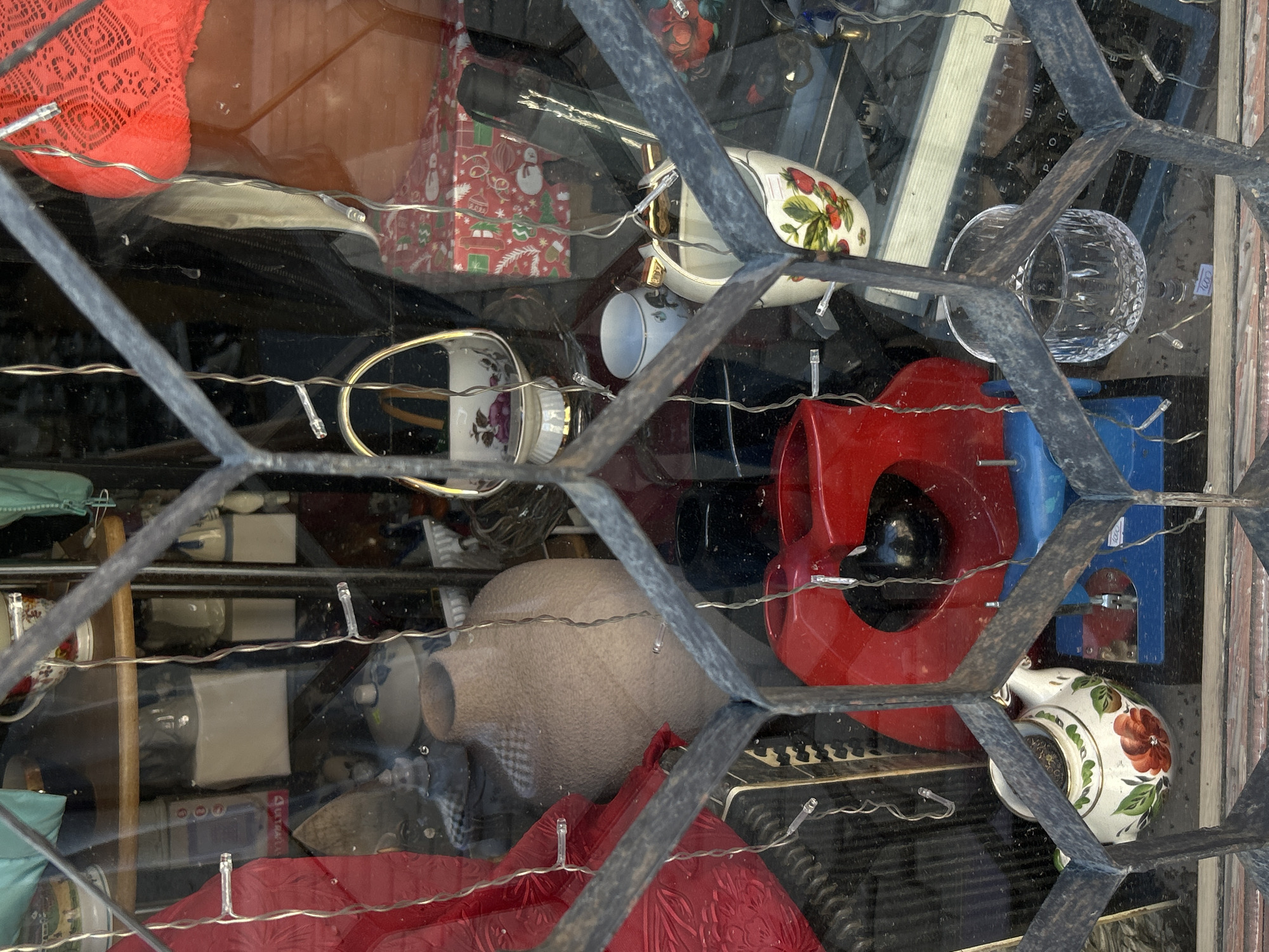 Through a dirty, diamond-patterned window pane, a cluttered interior is visible. The window frame is dark and weathered. Inside, a collection of miscellaneous objects includes a red knit item, a gift box with a Christmas pattern, a white cup with a strawberry design, a clear faceted glass, a red plastic horn-shaped item, a blue stapler, a beige vase, and a white teapot with a floral design. String lights are interspersed among the objects. The image appears to be taken in a cluttered retail space or storage area. The location is Saint Petersburg, Russia.