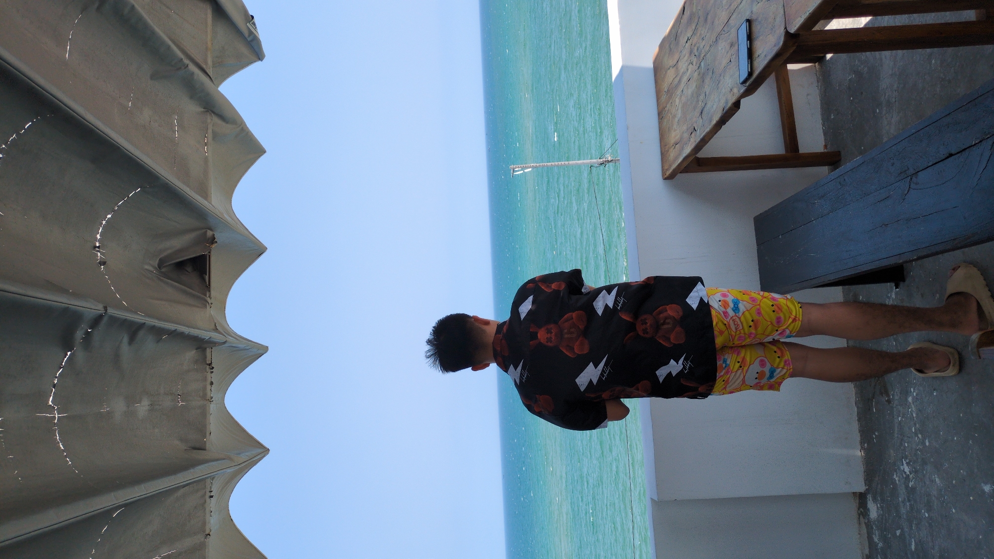 A person stands on a balcony overlooking the ocean in Phan Rang – Tháp Chàm, Vietnam. The person is wearing a black shirt with teddy bears and lightning bolts, and yellow shorts with cartoon characters. A wooden table and bench are to the person's right. A fabric canopy is on the left. The ocean and sky are in the background.