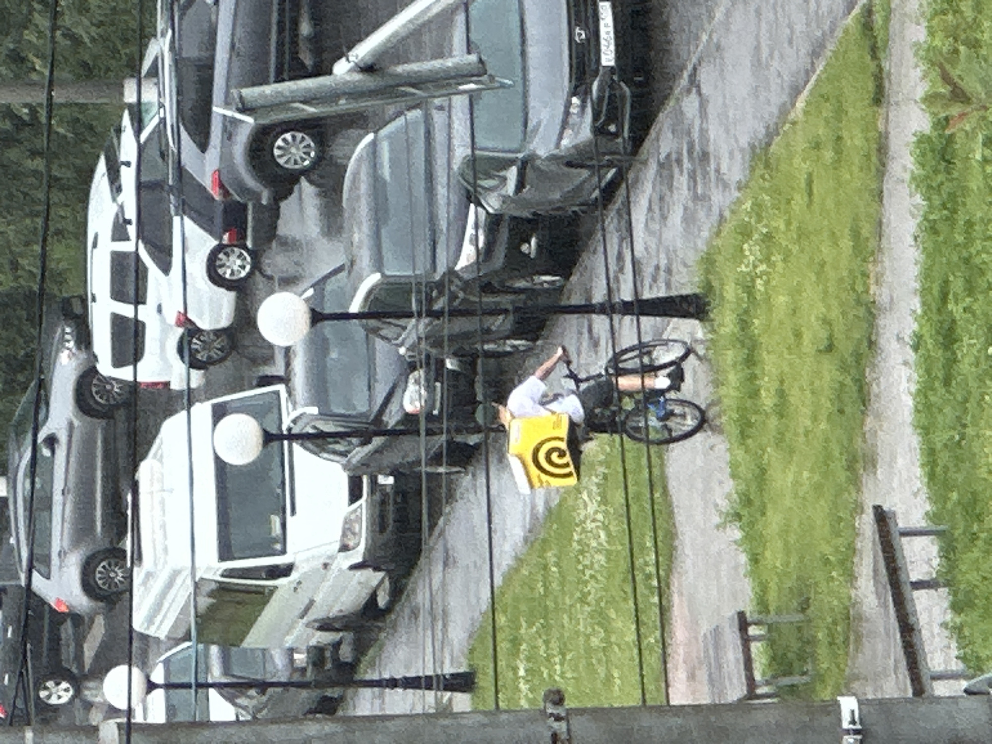 The image depicts a street scene in Podolsk, Russia, possibly on a rainy day. In the foreground, a person wearing a white shirt, dark shorts, and a dark baseball cap is riding a bicycle. They are carrying a bright yellow insulated delivery box with a spiral logo on their back. The bicycle has blue accents. To the left of the cyclist, there are two wooden benches on a grassy area. The ground appears wet, suggesting recent rain.
In the background, several vehicles are parked or driving on the wet asphalt road. These include a white van, a dark grey SUV, and other cars. The buildings and foliage in the distance are out of focus. The overall impression is a typical urban street scene with elements of a delivery service.
