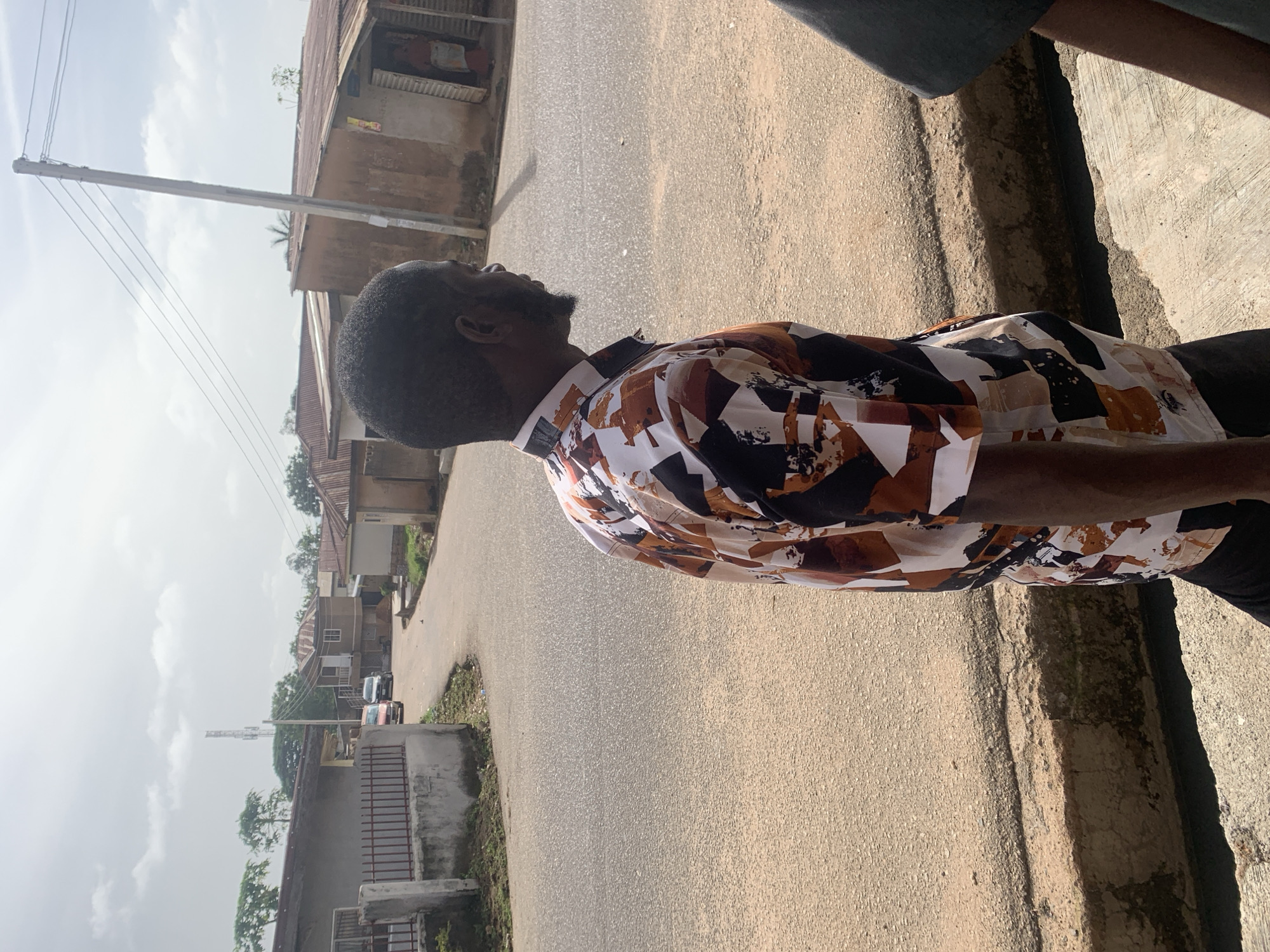 In the city of Jos, Nigeria, an individual wearing a colorful patterned shirt stands on a dirt path beside a paved road. The man, viewed from the side, has dark skin, a beard, and short dark hair. He is looking to the right. In the background, a variety of buildings with corrugated metal roofs are visible, along with trees and a distant cell tower. The sky is overcast with scattered clouds. The road curves gently, with vehicles parked in the distance and houses lining its sides. A concrete curb separates the path from the road.