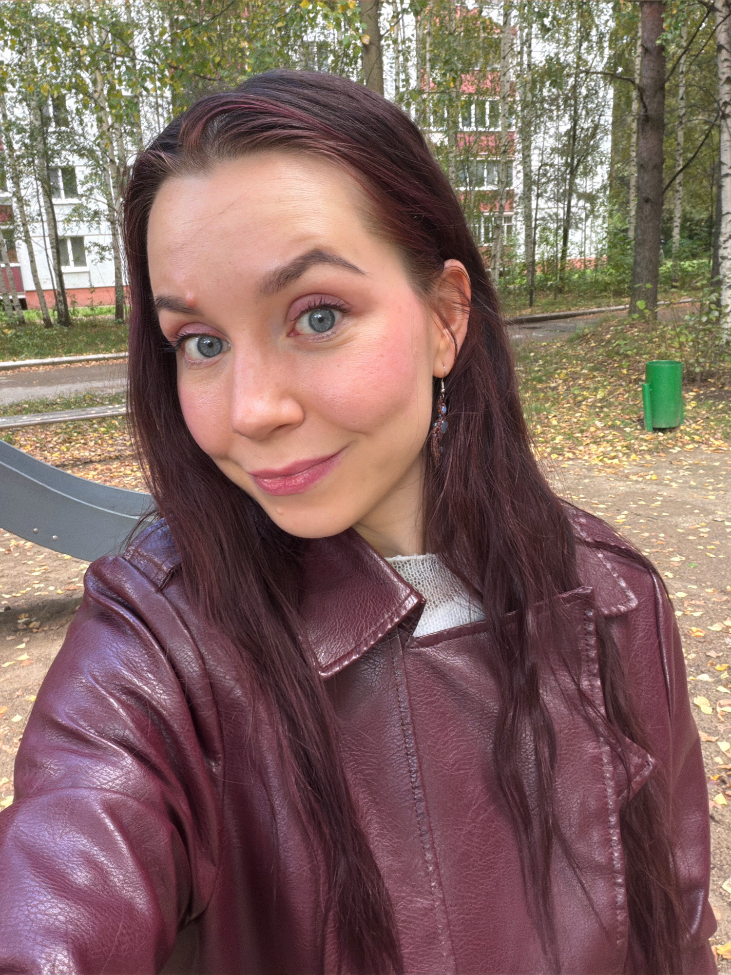 The image is a close-up, waist-up selfie of a young Caucasian woman with long, dark auburn hair. She is wearing a burgundy leather jacket over a light-colored sweater. Her eyes are blue and she is smiling softly. Her makeup includes pink blush and lip gloss. She is wearing a dangling earring in her visible ear. The background suggests an outdoor, urban park-like setting with trees, fallen leaves on the ground, a portion of a playground slide, and a residential building with windows in the distance. There is also a green trash receptacle visible. The overall atmosphere suggests an autumn day. The location is identified as a playground in the city of Chaikovsky, Russia.