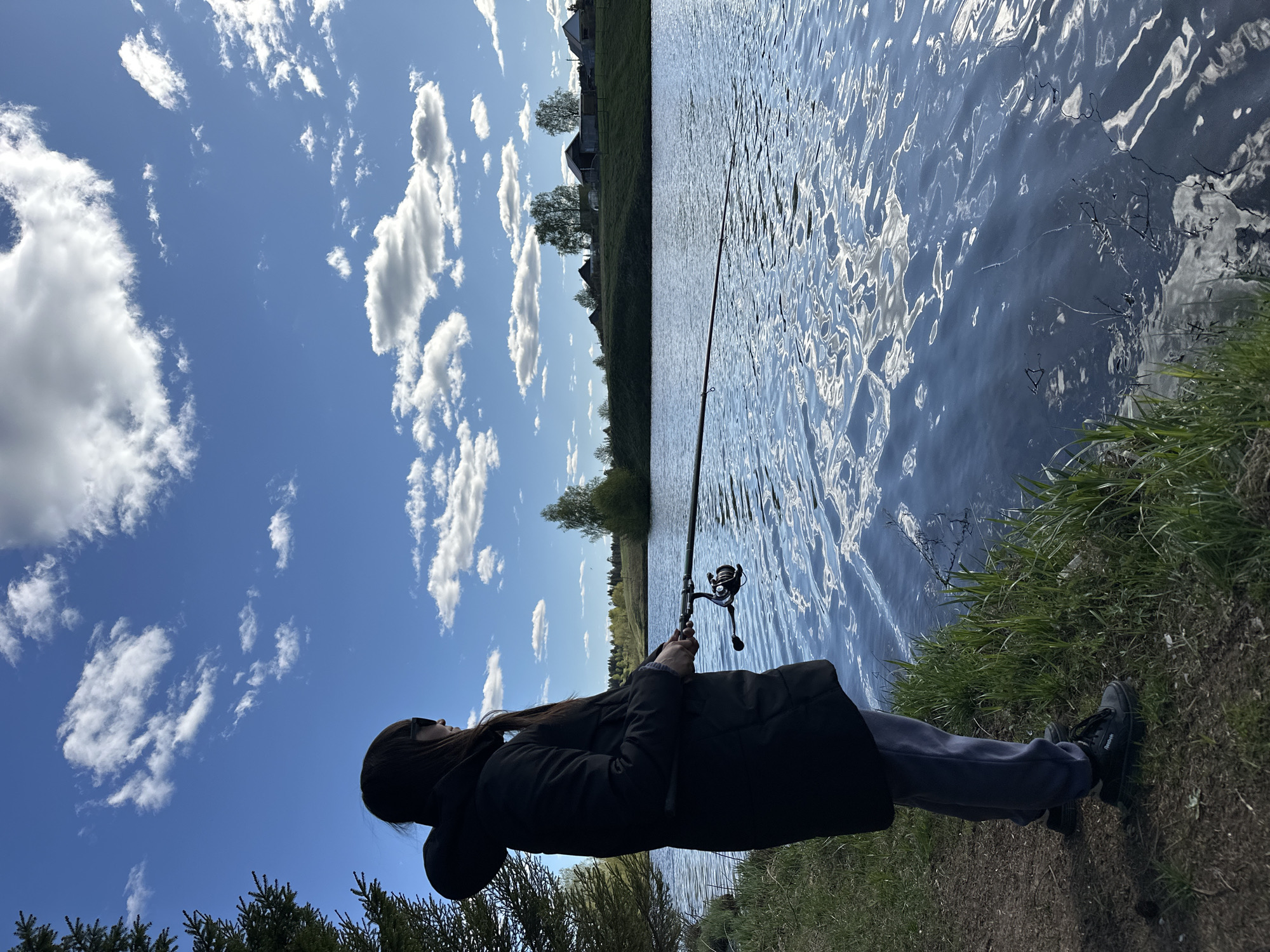 A person is fishing at the edge of a body of water, possibly a pond or lake, under a blue sky with scattered clouds. The person is wearing a dark coat and light-colored pants, holding a fishing rod with a reel. The edge of the water is grassy. In the distance, trees and buildings can be seen. The location is possibly near Buranovo, Russia.
