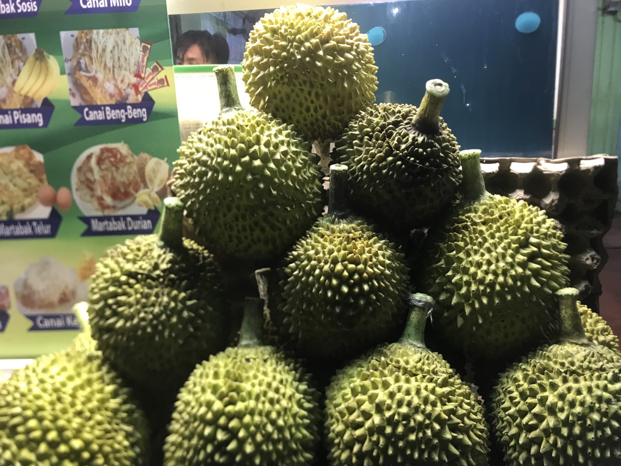 A stack of durian fruit is presented in a well-lit indoor setting, likely in Lhokseumawe, Indonesia. The pile consists of about a dozen spiky, green and yellowish durians of various sizes. In the background, a menu board displays images of different food items, including "Canai Pisang," "Canai Beng-Beng," "Martabak Telur," and "Martabak Durian," suggesting this is a food vendor specializing in local cuisine. Behind the fruits, there's a partially visible individual with dark hair looking in the direction of the fruits. The overall scene conveys a sense of a local food market or restaurant.