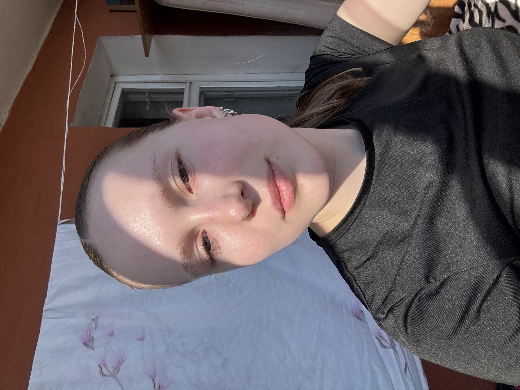 A young woman is lying down, possibly outdoors, with her head and upper body visible. She has fair skin and light brown hair, wearing a black top. Her face is illuminated by strong sunlight. She has jewelry visible in her ear. A white sheet with a floral pattern is beneath her. The environment suggests an outdoor setting with a window visible in the background and architectural elements. There is no specific information that indicates the location being in Chaikovsky, Russia.