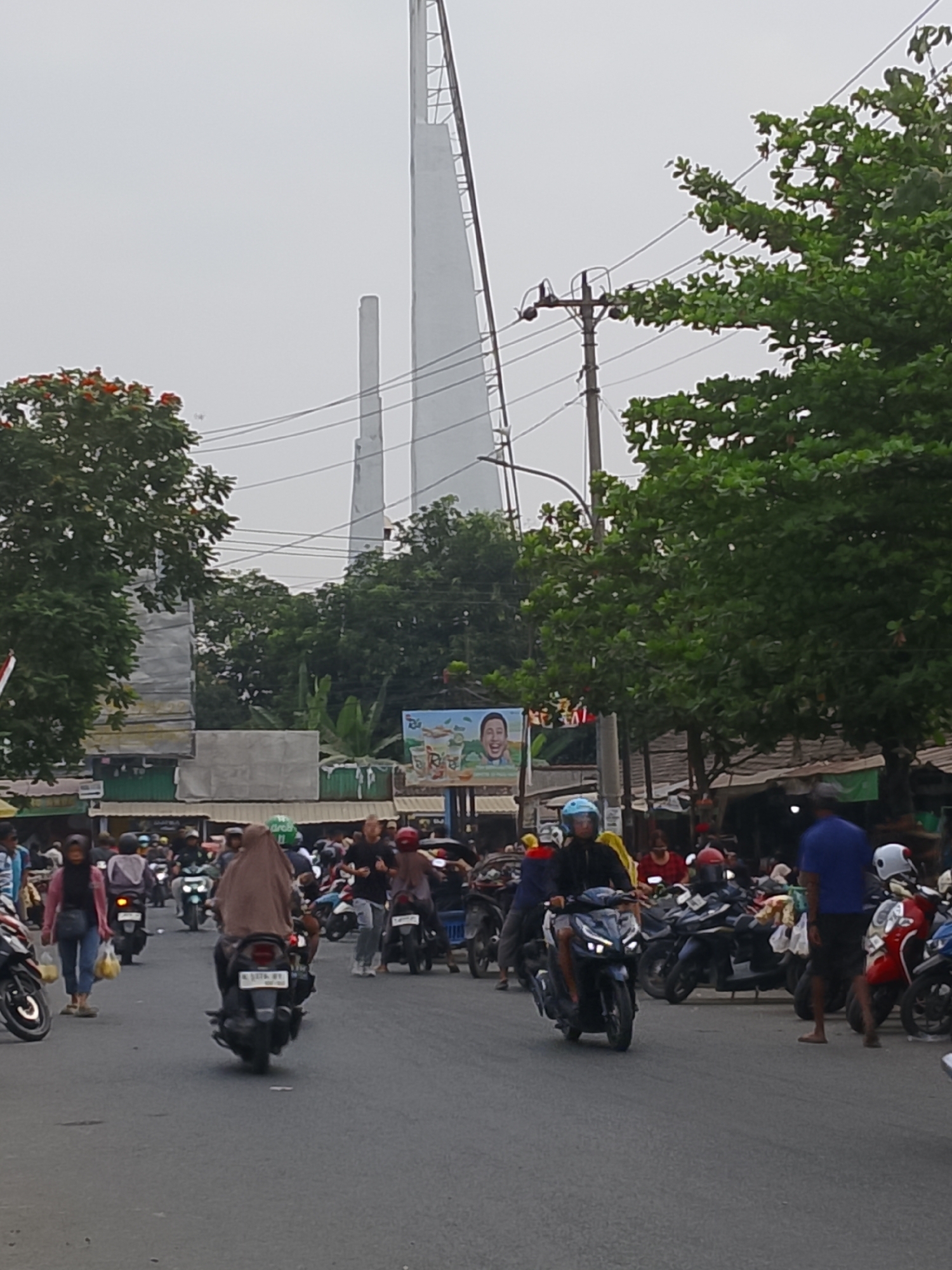 The image depicts a street scene in Ngelo, Indonesia, likely during daylight hours, with an overcast sky. The foreground is dominated by a paved road filled with numerous motorcycles and scooters, many of which are being ridden by people wearing helmets. A diverse group of individuals, including men and women, are visible, some walking and others on motorcycles. Several people are carrying shopping bags.
In the background, there are buildings with awnings, suggesting a commercial or market area. Trees with green foliage and some with red flowers are present on the sides of the road and behind the buildings. Prominently in the distance, two tall, white, spire-like structures are visible, their tops extending into the hazy sky. One of these structures appears to be a series of interconnected poles, while the other is a solid, tapered column. Electrical wires and poles are also part of the urban infrastructure. A billboard with an image of a smiling man and text is positioned between two of the buildings. The overall atmosphere suggests a bustling urban environment.