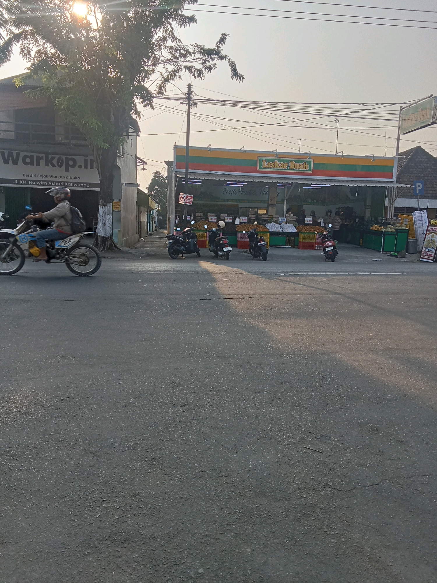 The image depicts a street scene in Kaliwungu, Indonesia. A person is riding a dirt bike from left to right across the foreground. To the right of the dirt bike rider, a building with a sign that reads "Warkop.in" is visible, with text below indicating "Jl. KH. Hasyim Ashari Kaliwungu." Further to the right, a brightly colored storefront with horizontal stripes in red, yellow, and green displays the name "Laskar Buah." This establishment appears to be a fruit stand or market, with various fruits, baskets, and people visible inside and in front of it. Several motorcycles are parked in front of the Laskar Buah establishment. The environment suggests a commercial area with ongoing activity, possibly in the late afternoon given the lighting.