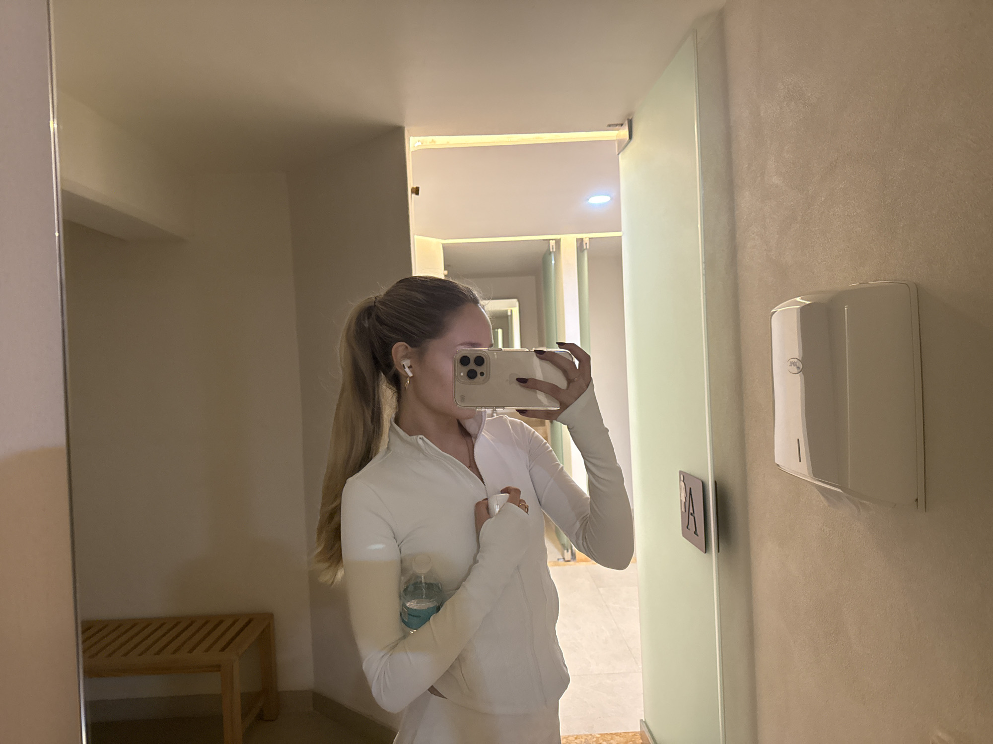 A person with long blonde hair tied in a ponytail, wearing a white long-sleeved athletic top and white wireless earbuds, is captured in a mirror selfie. Their face is obscured by a light-colored smartphone, held with both hands, positioned to take the photo. A clear plastic water bottle with a blue label is held in their left arm. The background, visible in the mirror's reflection, depicts an indoor changing or locker room environment. To the left, a light wooden slatted bench is visible. To the right, a white wall-mounted dispenser with faint branding is affixed to a textured light beige wall. Adjacent to the dispenser, a dark sign with a white "A" and an indistinct symbol is mounted on a light green partition or door frame. Beyond this, a hallway with further light green partitions and overhead lighting is visible. The specific location within Mexico City, Mexico, cannot be determined from the visual information provided in the image.
