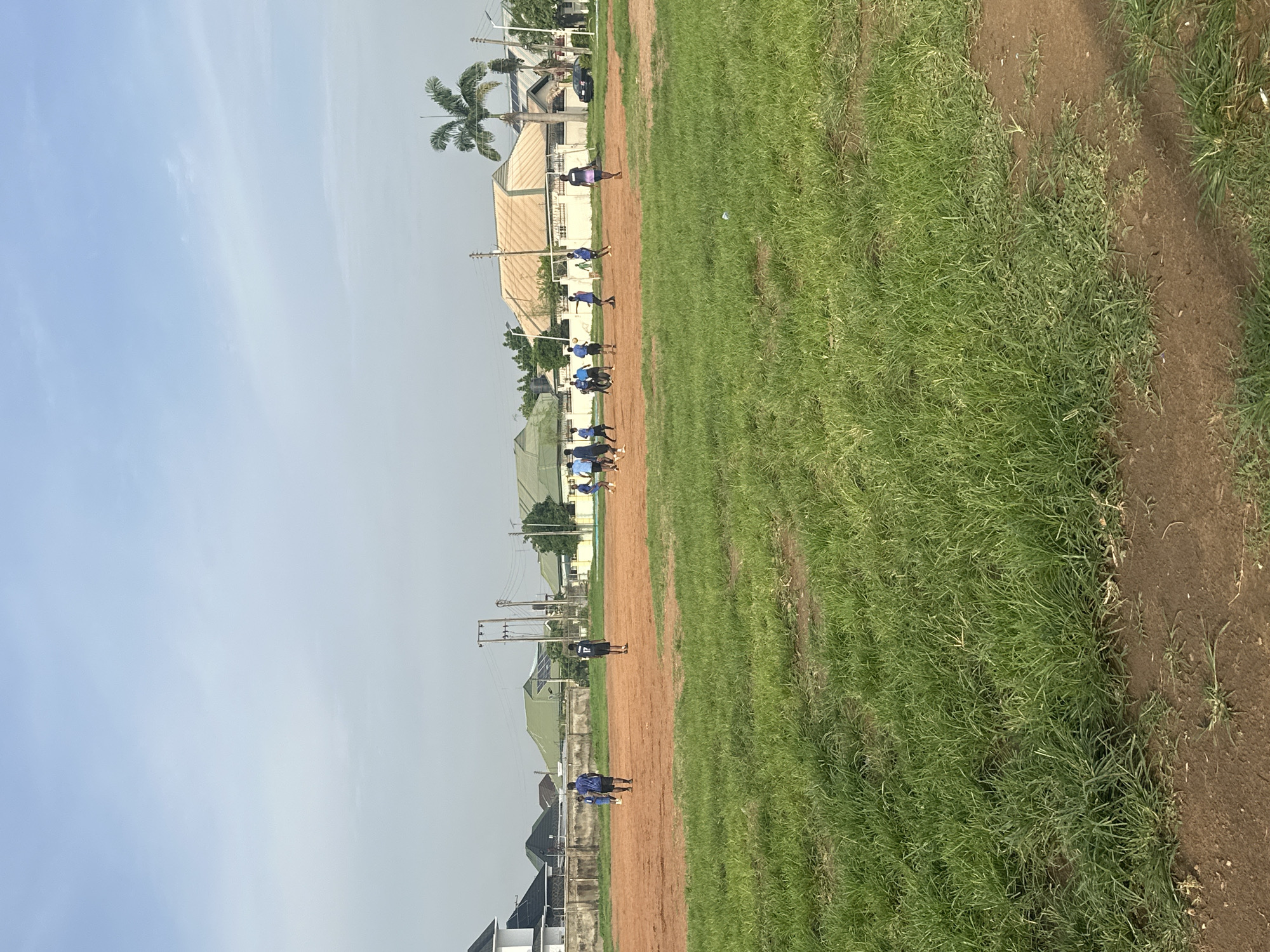 This media file captures a view of a dusty sports field with patches of green grass, likely located in Abuja, Nigeria. The image is oriented vertically, with the sky occupying the upper left portion and the field extending to the right. A dirt track runs diagonally across the middle of the frame, separating the grassy area from a line of buildings and a palm tree on the left.
Several people, appearing to be young individuals, are spread out along the dirt track. They are wearing dark blue or purple athletic wear and seem to be engaged in some form of outdoor activity, possibly training or a race, given their scattered positions and the setting. They are small figures in the frame, indicating they are some distance away.
In the background, various structures are visible, including houses with light-colored roofs and some trees. Electrical poles and wires are also present, suggesting an inhabited area. The sky is a light blue with faint clouds, indicating it is likely daytime, possibly late morning or early afternoon. The overall atmosphere appears to be casual and active, with the focus on the individuals training in the outdoor environment. There is no visible text in the image.
