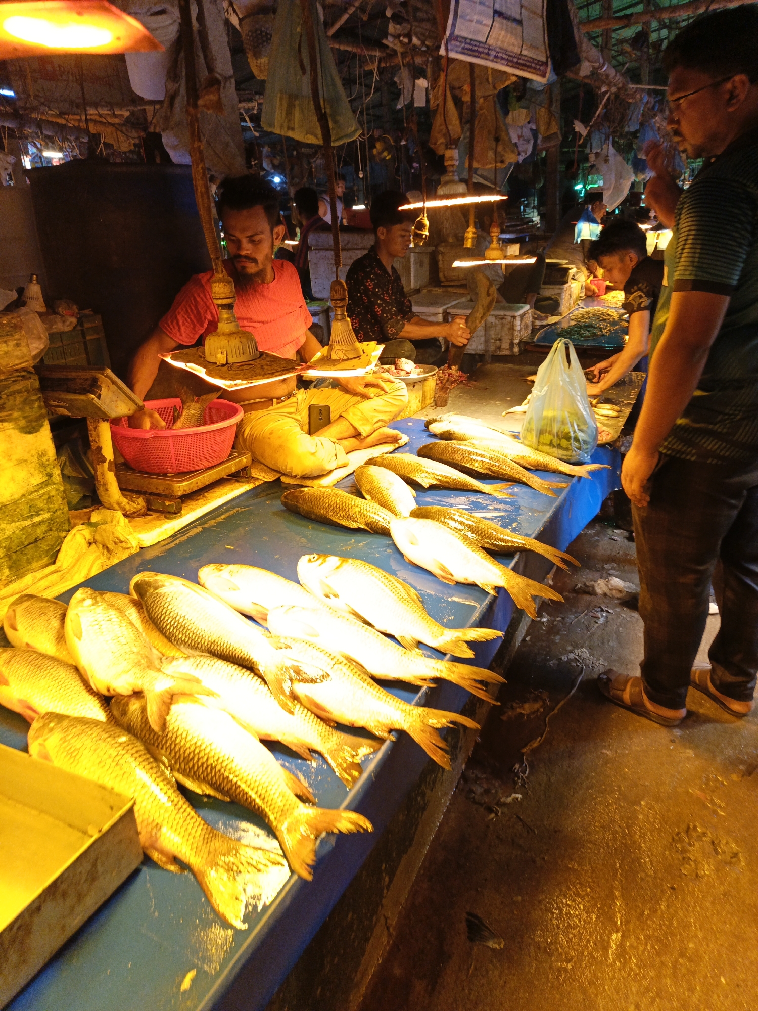 The image depicts a busy fish market scene, likely in Dhaka, Bangladesh. A vendor in a red shirt is weighing fish using a scale with a red basket on a blue table. Numerous silver-colored fish are lined up for sale on the table. Customers are interacting with the vendor. The lighting is warm, with lamps hanging above the stalls. The environment appears cluttered, with various items hanging from the ceiling, adding to the chaotic atmosphere of the market. The floor is wet, reflecting the light.