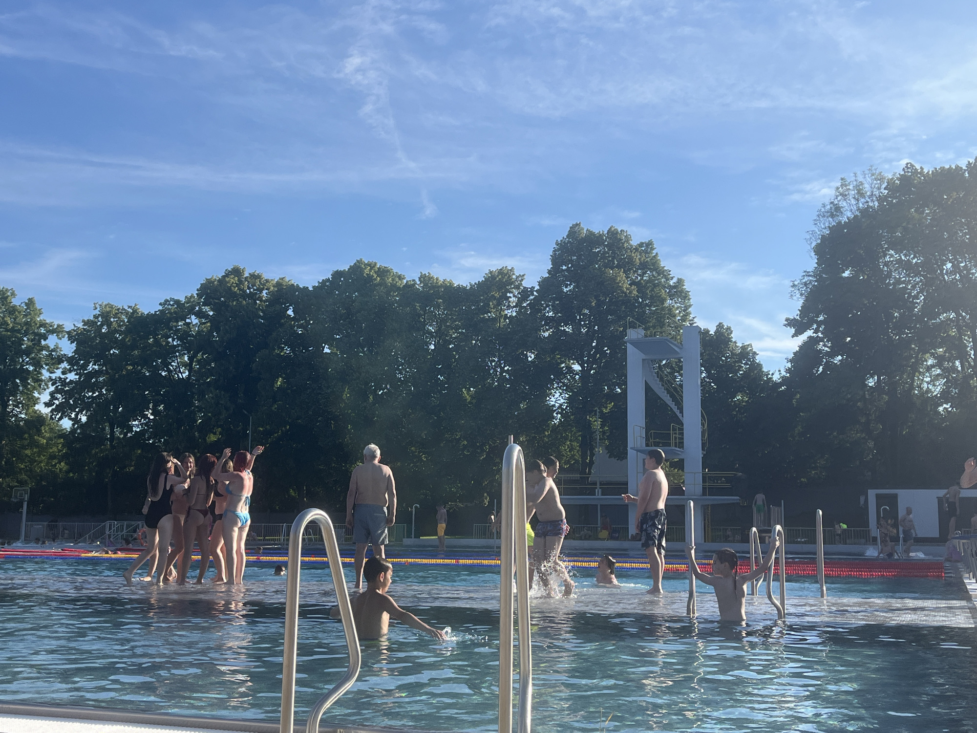 A group of people are enjoying a sunny day at an outdoor swimming pool. The water is clear and blue, reflecting the bright sky above. Several individuals are in the water, some swimming, others standing and socializing. In the foreground, two metal railings rise out of the water, part of the pool's entry and exit system.
On the left side of the pool, a group of young women in swimsuits are gathered, laughing and interacting. Further back, a man with a grey mustache stands near the edge of the pool, wearing swim trunks. Several other people are visible in the background, some in the water and others walking around the pool area.
A diving platform with multiple levels stands prominently in the background, suggesting a dedicated diving area within the pool complex. Lush green trees line the perimeter of the pool, providing shade and a natural backdrop. The sky is a clear blue with a few wispy clouds, indicating fair weather. The overall atmosphere appears relaxed and cheerful, with people enjoying recreational activities on a warm day. The sun casts a bright light, suggesting it is daytime, likely late morning or afternoon. No visible text is present in the image.