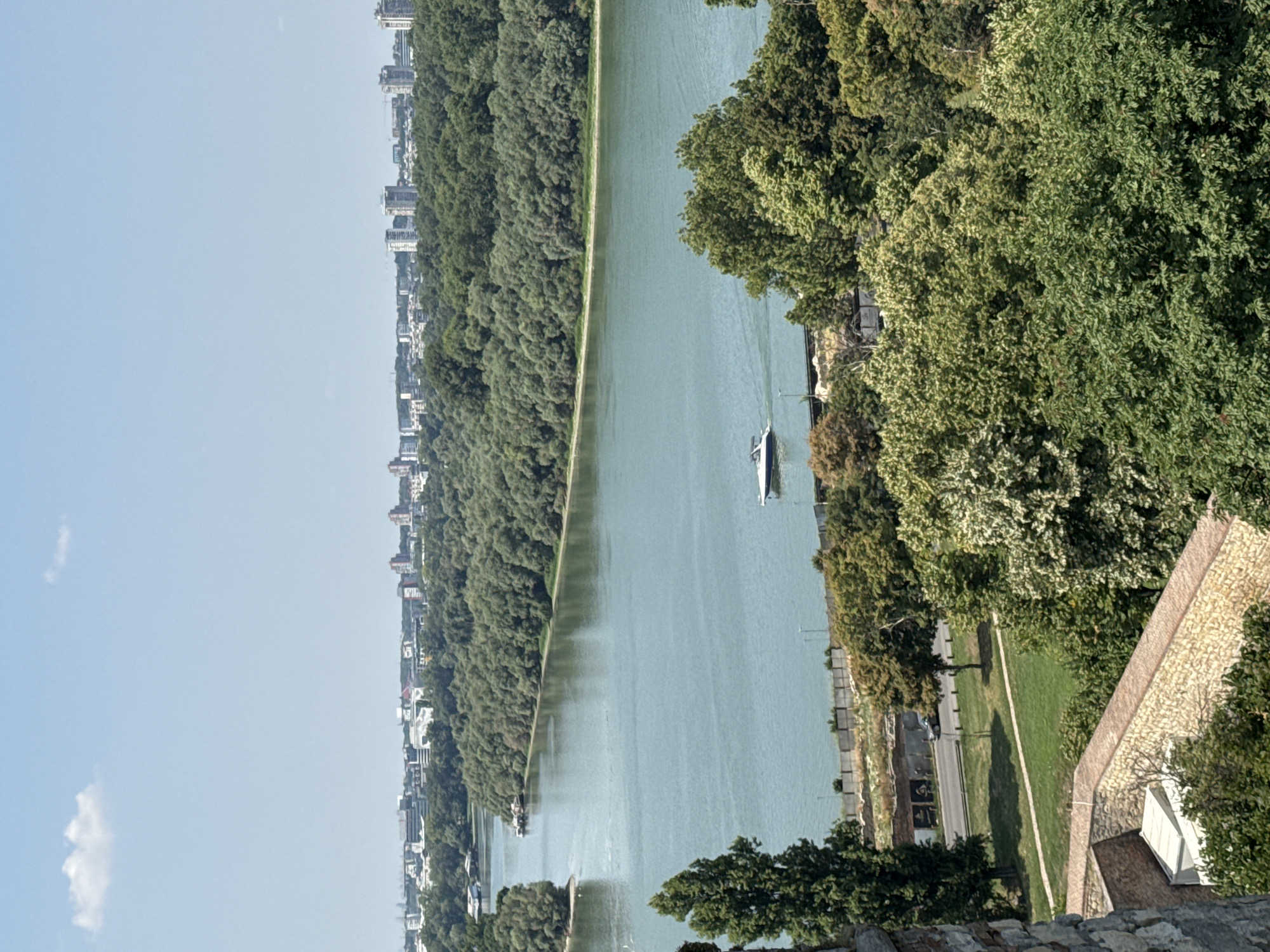 The image shows a wide, tranquil river with a clear blue sky overhead. A small white motorboat cruises on the water, leaving a gentle wake. The riverbanks are densely wooded with lush green trees, creating a natural, serene backdrop. In the distance, a cityscape with tall buildings is visible, suggesting an urban environment adjacent to the natural landscape. The foreground features elements of a stone structure, possibly a fortification, with greenery interspersed. The overall impression is one of a peaceful day in a city where nature and urban development coexist harmoniously. This view is characteristic of Belgrade, Serbia, overlooking the confluence of the Sava and Danube rivers.