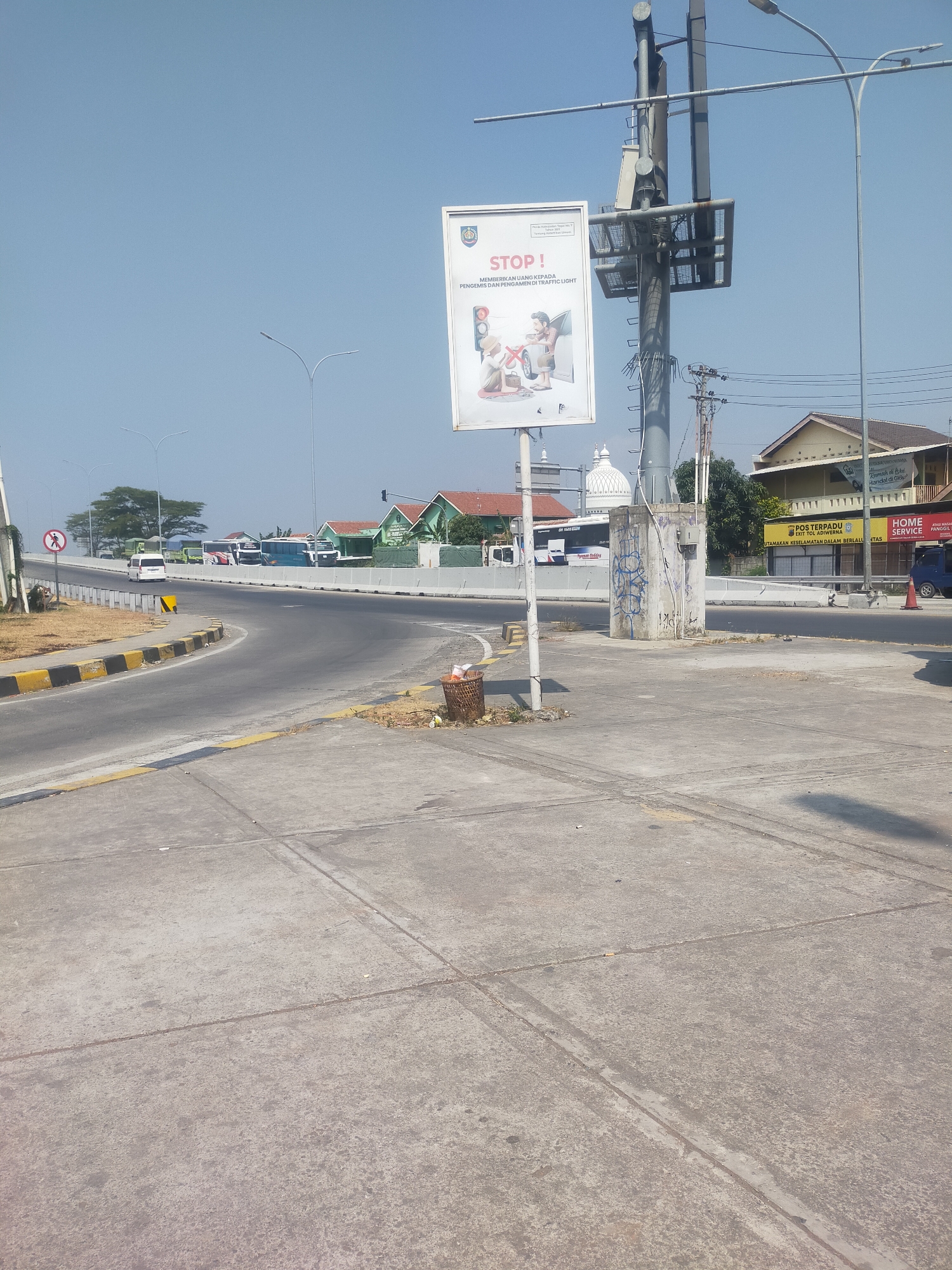 The image depicts a street scene in Lemahduwur, Indonesia, on a clear, sunny day. In the foreground, a concrete pedestrian area is visible, with distinct seams in the pavement. A metal trash bin, overflowing with refuse, sits to the right of the pedestrian area. Adjacent to this is a busy intersection with a road curving to the left.

A large billboard prominently displays a message with the word "STOP!" in red letters. Below this, the text, "MEMBERIKAN UANG KEPADA PENGEMIS DAN PENGAMEN TRAFFIC LIGHT" is visible, translating to "GIVING MONEY TO BEGGARS AND TRAFFIC LIGHT VENDORS." The graphic on the billboard illustrates this, showing two figures, one with coins and the other a beggar, with a traffic light symbol superimposed.

To the right of the billboard, a tall, industrial pole with various equipment and wiring rises towards the sky. Further down the street, several buildings with tiled roofs are visible, including what appears to be a domed building, possibly a mosque. Multiple buses and a white van are parked or in motion on the road. A traffic signal can be seen in the distance. The environment suggests a semi-urban or commercial area with a considerable amount of vehicular traffic.