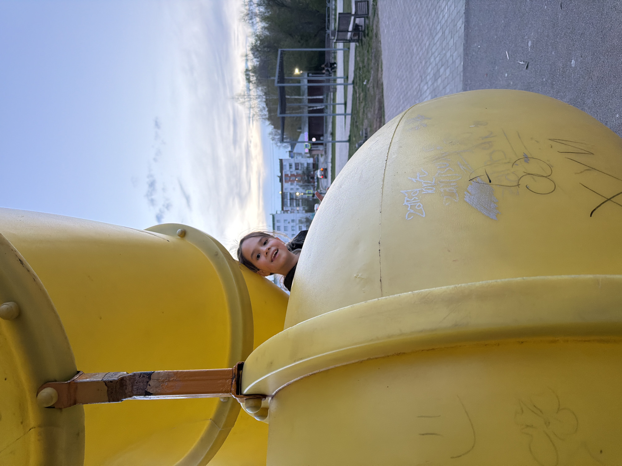 A young girl is peering out from between two yellow playground structures, possibly parts of a slide or climbing apparatus. The structures appear to be made of plastic or a similar material. There is graffiti on the structure to the right. The girl is smiling and looking directly at the camera. In the background, there is a park area with benches, metal structures, buildings, and trees. The sky is visible and it appears to be either early morning or late evening. Based on the surrounding environment, the location is Chaikovsky, Russia.