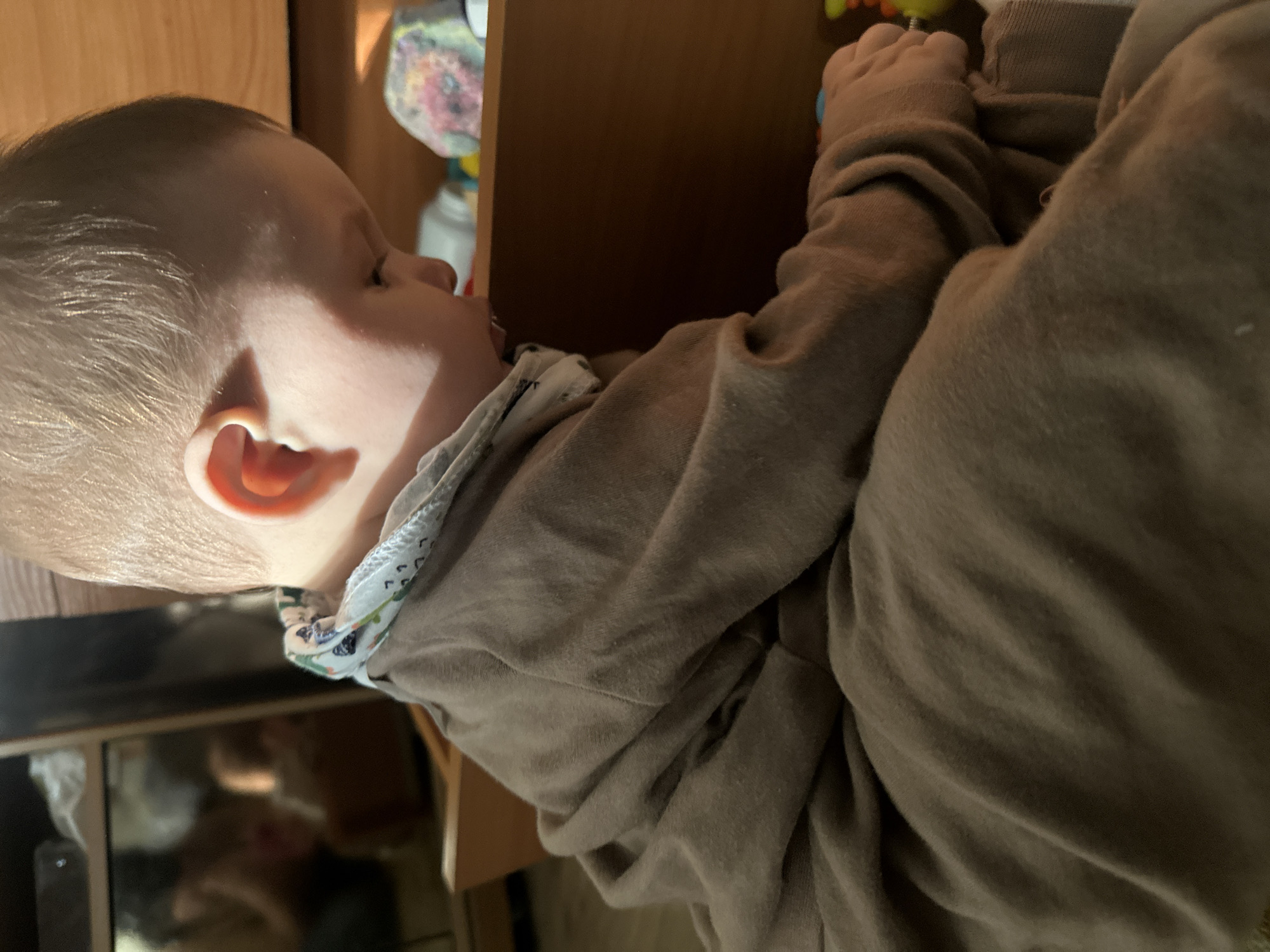 A close-up, tilted, indoor shot shows a baby in a brown outfit lying next to a wooden surface. The baby's face is turned to the right with eyes closed. Their ear is visible, and they wear a bib or collar with a light-colored pattern. The setting includes a brown wooden structure and other objects, possibly toys, in the background. A dark reflection appears at the bottom of the image. The location is Unknown, Unknown.
