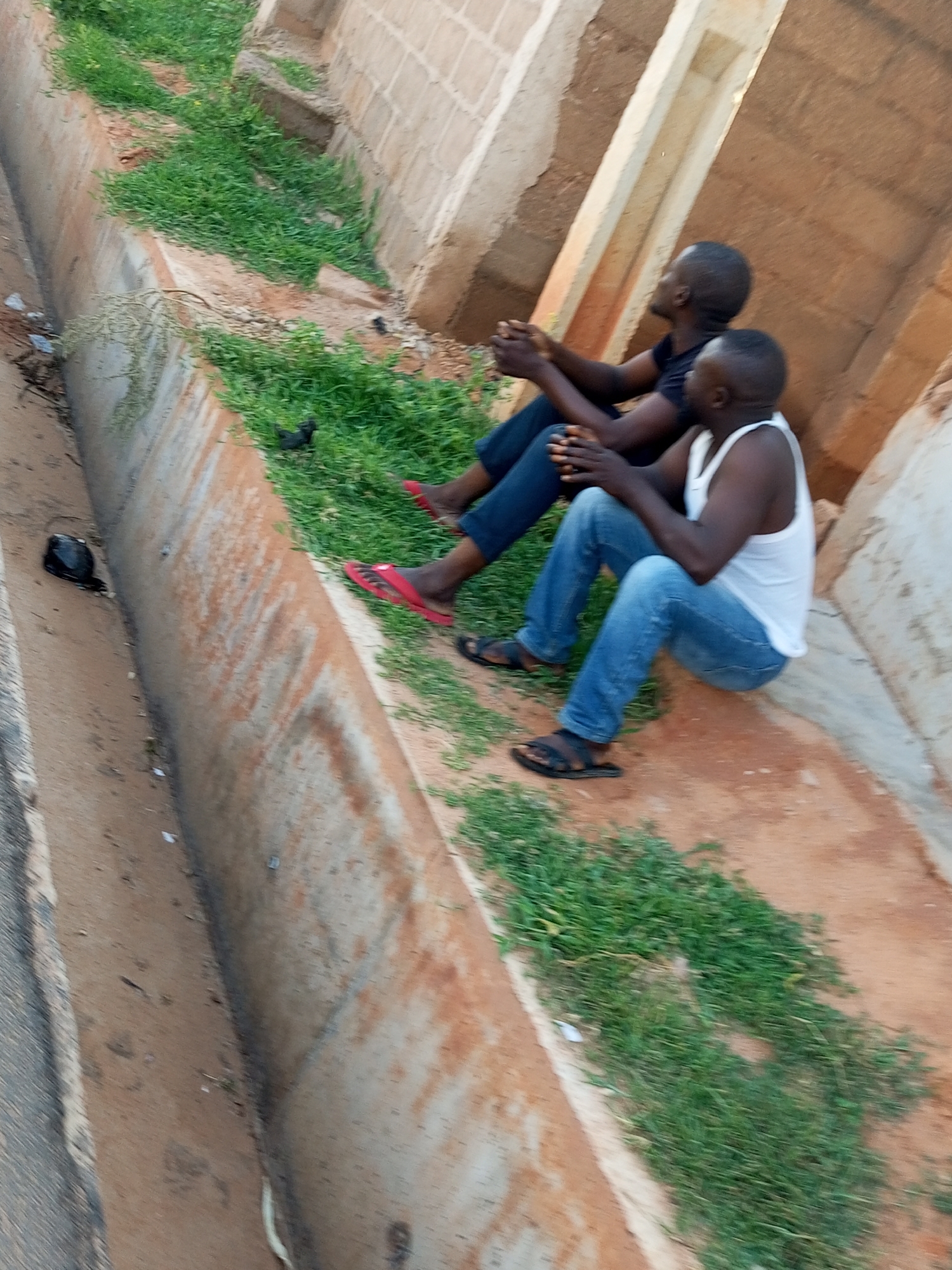 Two men are sitting on the edge of a concrete drainage ditch, their backs to the camera, in Gombe, Nigeria. The man on the left is wearing a dark t-shirt and blue jeans, while the man on the right is wearing a white tank top and blue jeans. Both men are wearing sandals. The drainage ditch is overgrown with grass and has some loose dirt and debris on the sides. The background consists of a rough concrete wall and a paved surface. The lighting suggests it is daytime, possibly late afternoon, with shadows cast on the wall. A black object, possibly a shoe or a bag, is on the ground near the drainage ditch.
