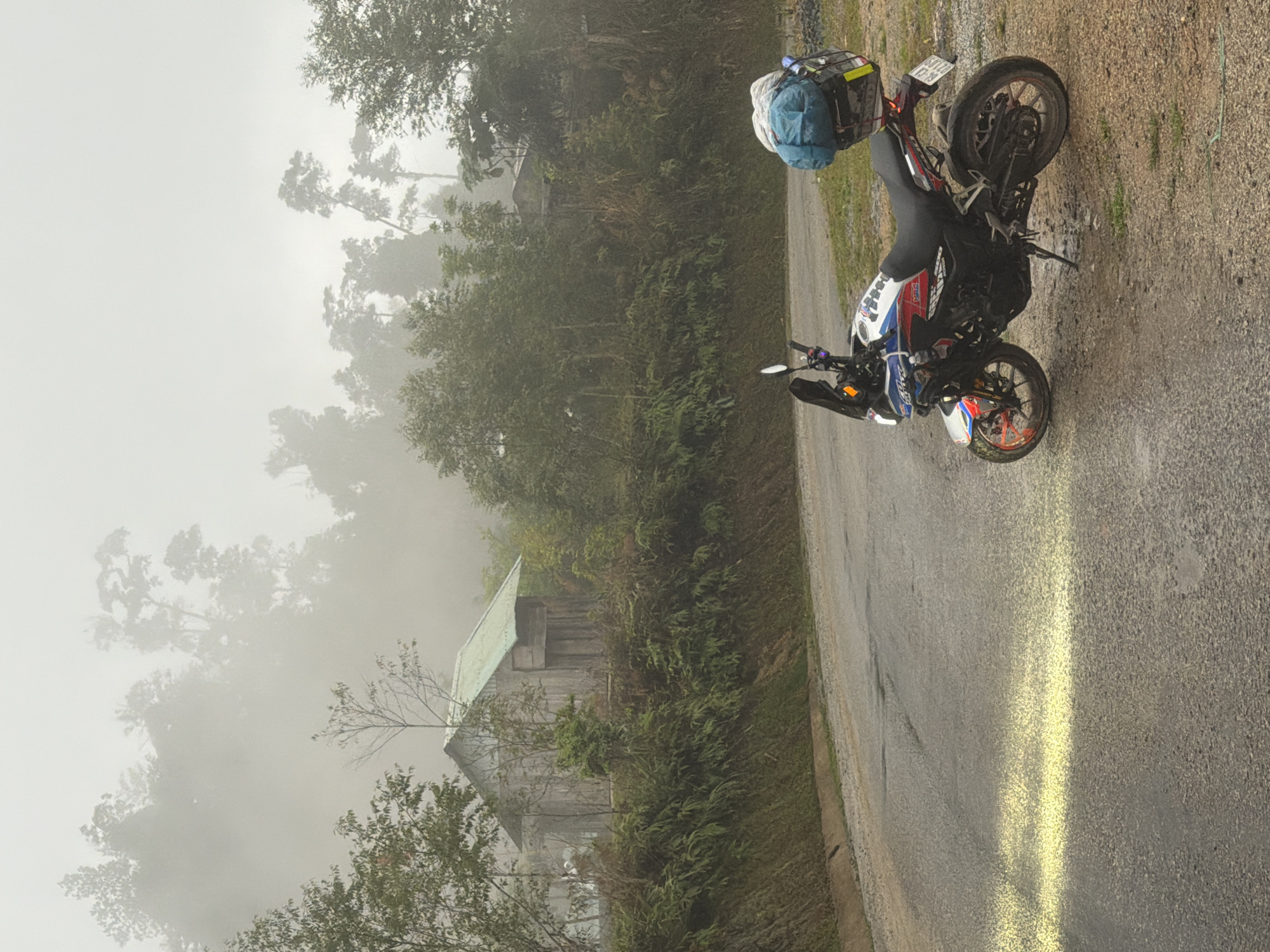 The image shows a motorcycle parked on a paved road alongside a hillside obscured by fog. The motorcycle is loaded with a blue duffel bag and a metal container, possibly for storage. The road surface appears wet. In the background, a weathered wooden structure with a metal roof is visible amidst dense foliage and trees shrouded in mist. The location is suspected to be in the city of Đà Nẵng, Vietnam. No people are visible in the frame.